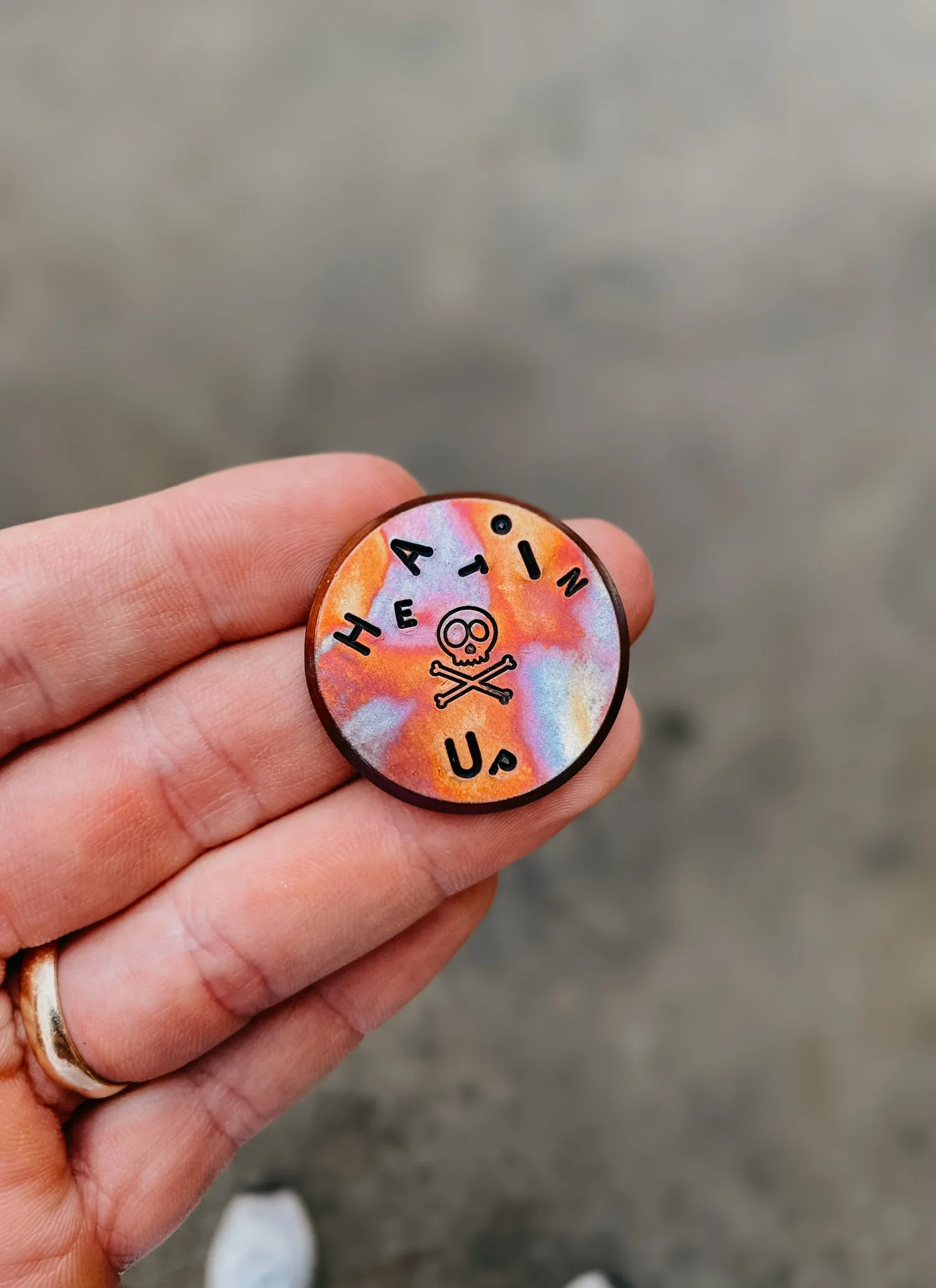 A hand holding a circular pin with a colorful, tie-dye background. The pin has the words "HEAVEN" and "USA" arranged around a small skull and crossbones design.