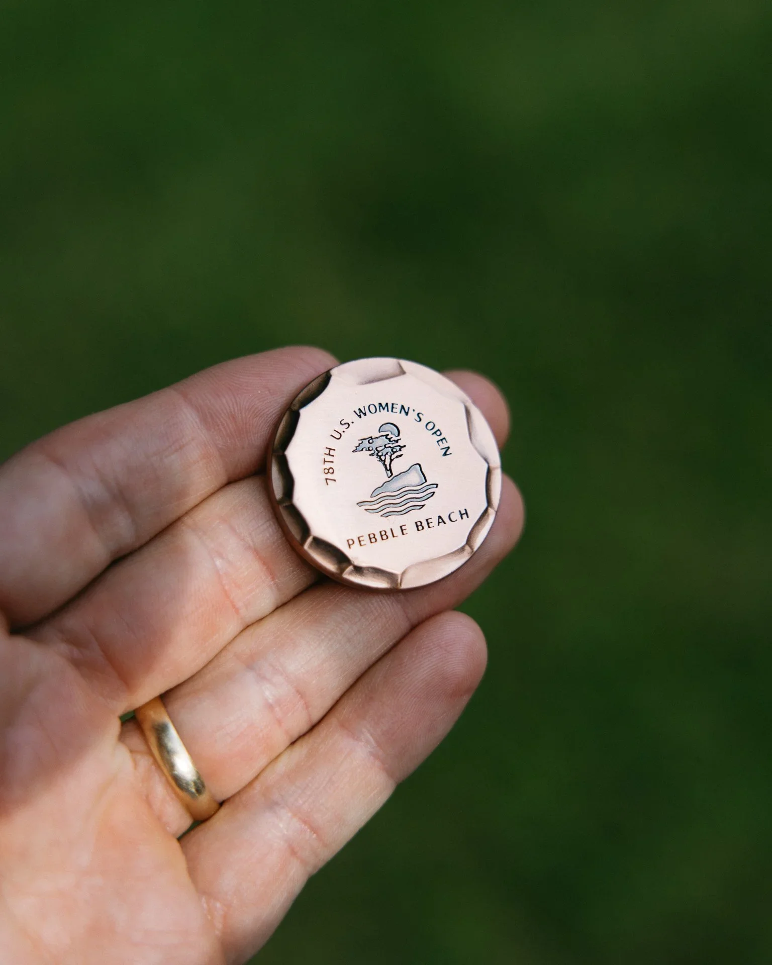 Hand holding a coin with text about the 78th U.S. Women's Open at Pebble Beach and a logo of a pine tree, water, and a cloud.