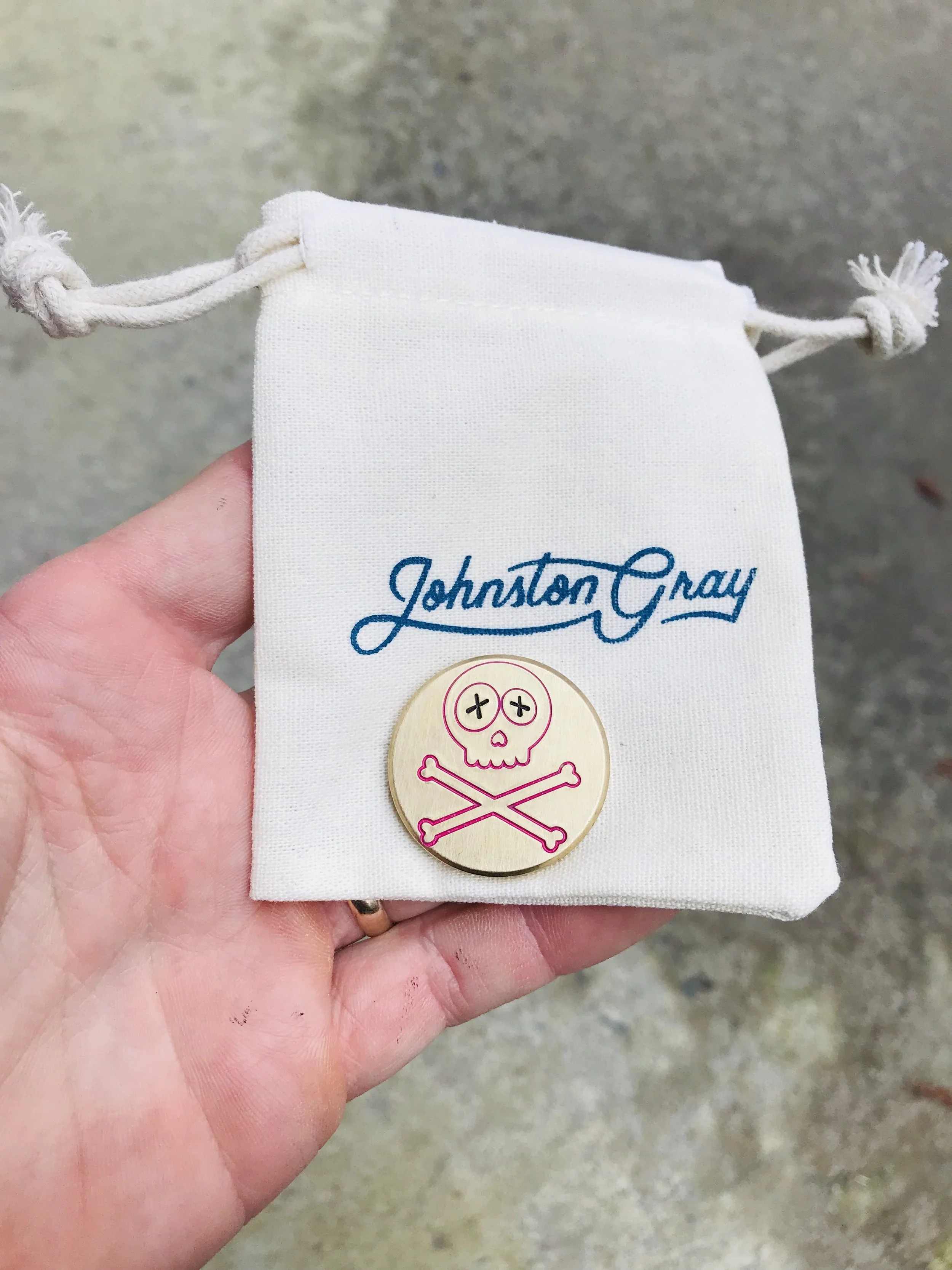 A hand holding a white drawstring pouch with the blue embroidered name 'Johnston Gray' and a round pin featuring a pink skull and crossbones design.
