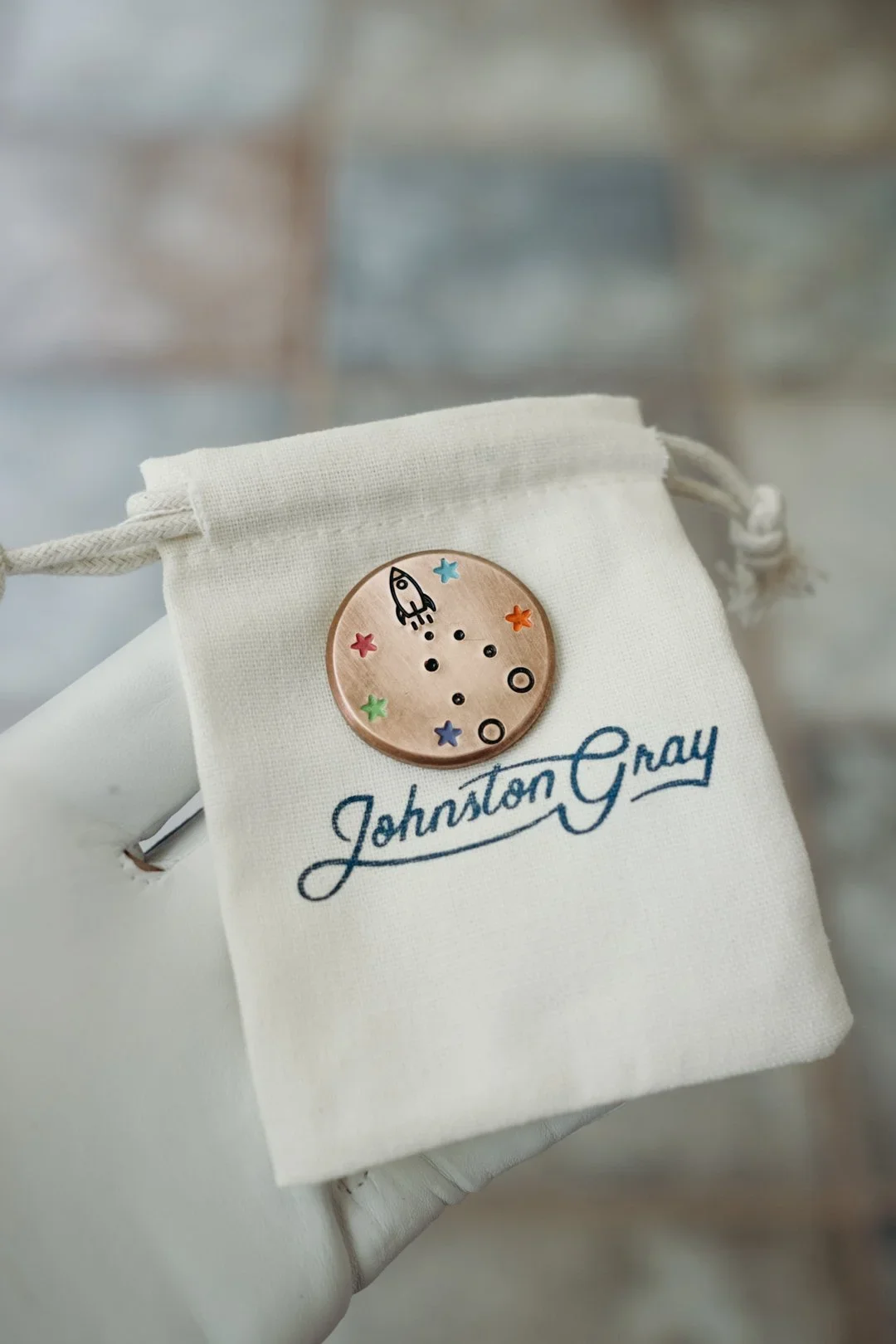 A white cloth bag with 'Johnston Gray' embroidered in blue, featuring a decorative metal pin of a moon with colorful stars and space-themed illustrations.