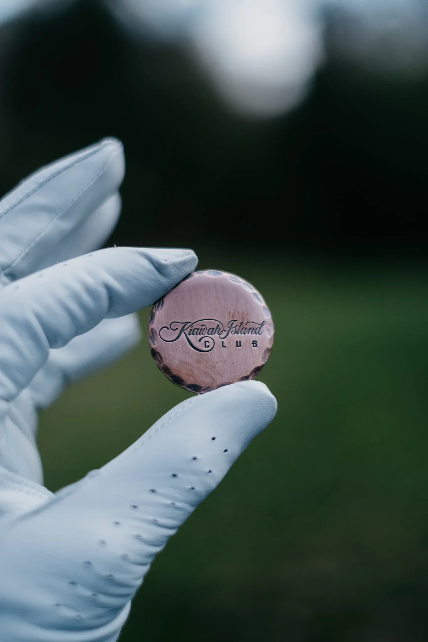 A person wearing white gloves holding a small, round, shiny coin with an inscription reading 'Kiawah Island Club' against a blurred green background.