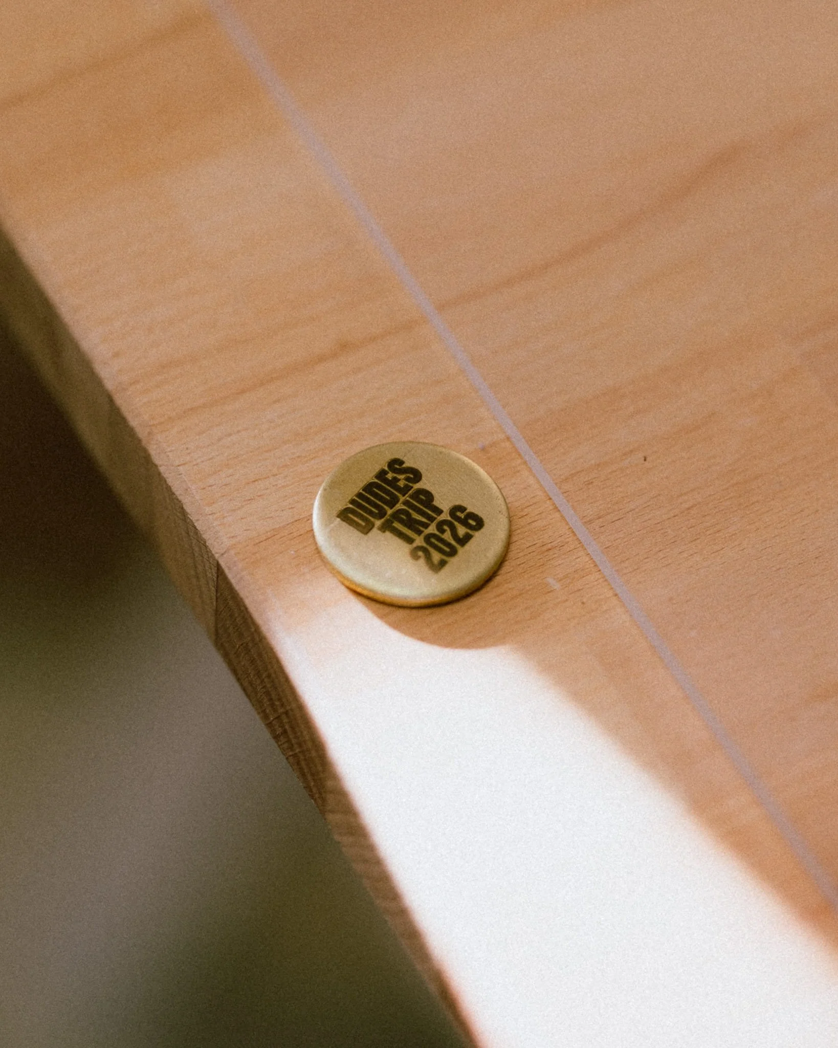 A close-up of a gold-colored circular badge on a wooden surface, with the text 'Dudes Trap 2026' printed on it.