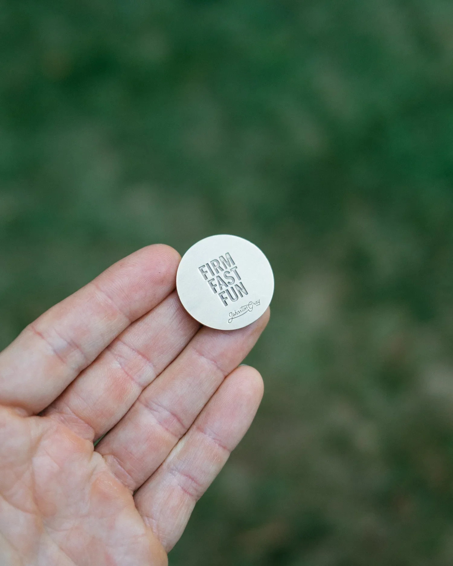 A hand holding a round white pin with embossed text that reads 'FIRM FAST FUN' and a signature underneath, against a blurred green background.