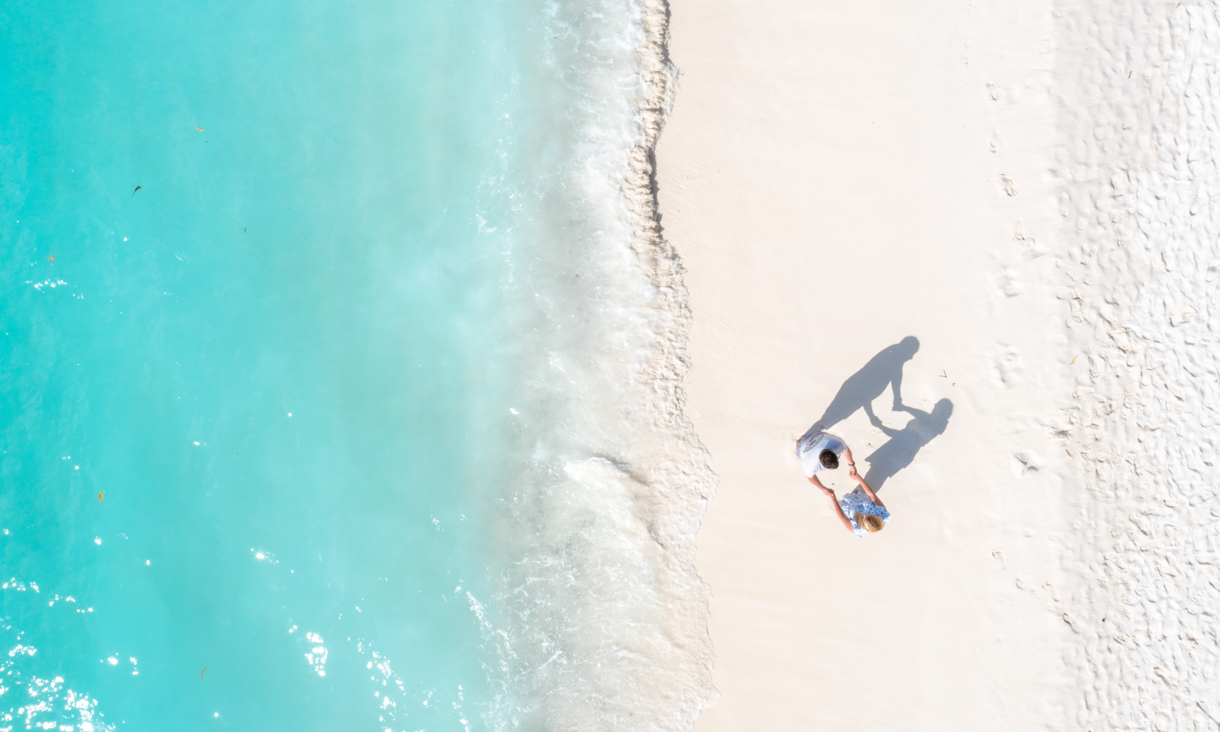 An aerial view of two people standing on white sandy beach near light blue ocean waves.
