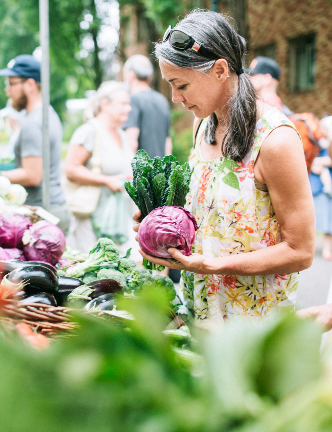 Woman at a farmer's market holding a purple cabbage and kale with other vegetables displayed on the table, with blurred people in the background.