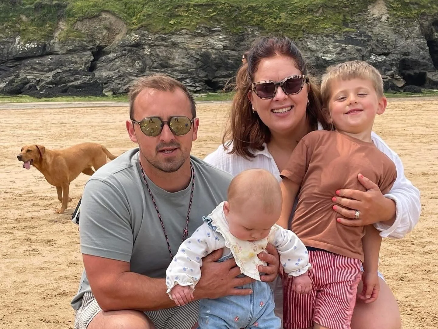 A family of four, including a man, woman, and two young children, smiling and posing for a photo on a sandy beach with a rocky cliff and a brown dog in the background.