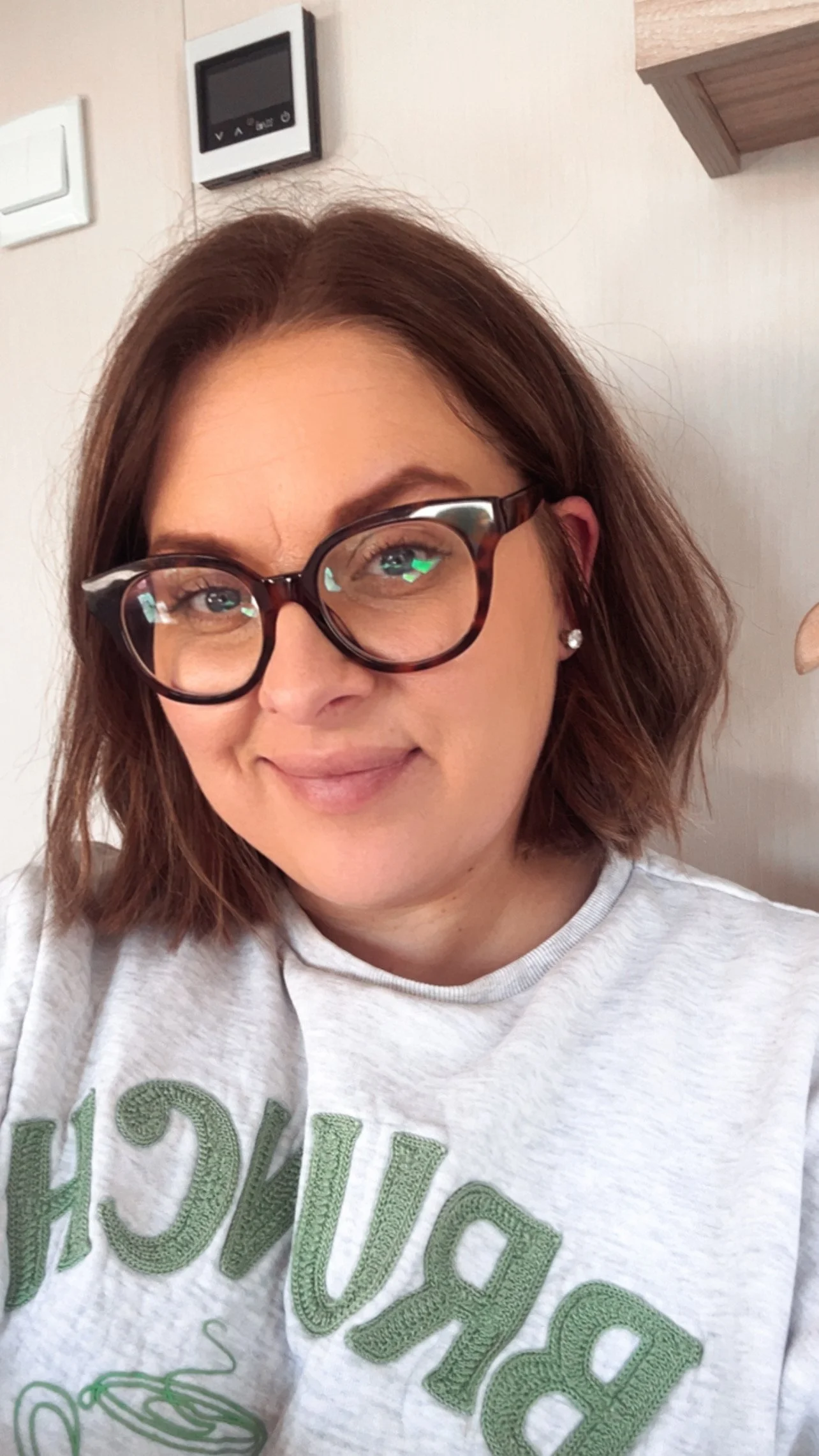 A woman with short, wavy brown hair wearing glasses and a light gray t-shirt with green embroidered text. She is smiling and sitting indoors with a beige wall and a shelf in the background.