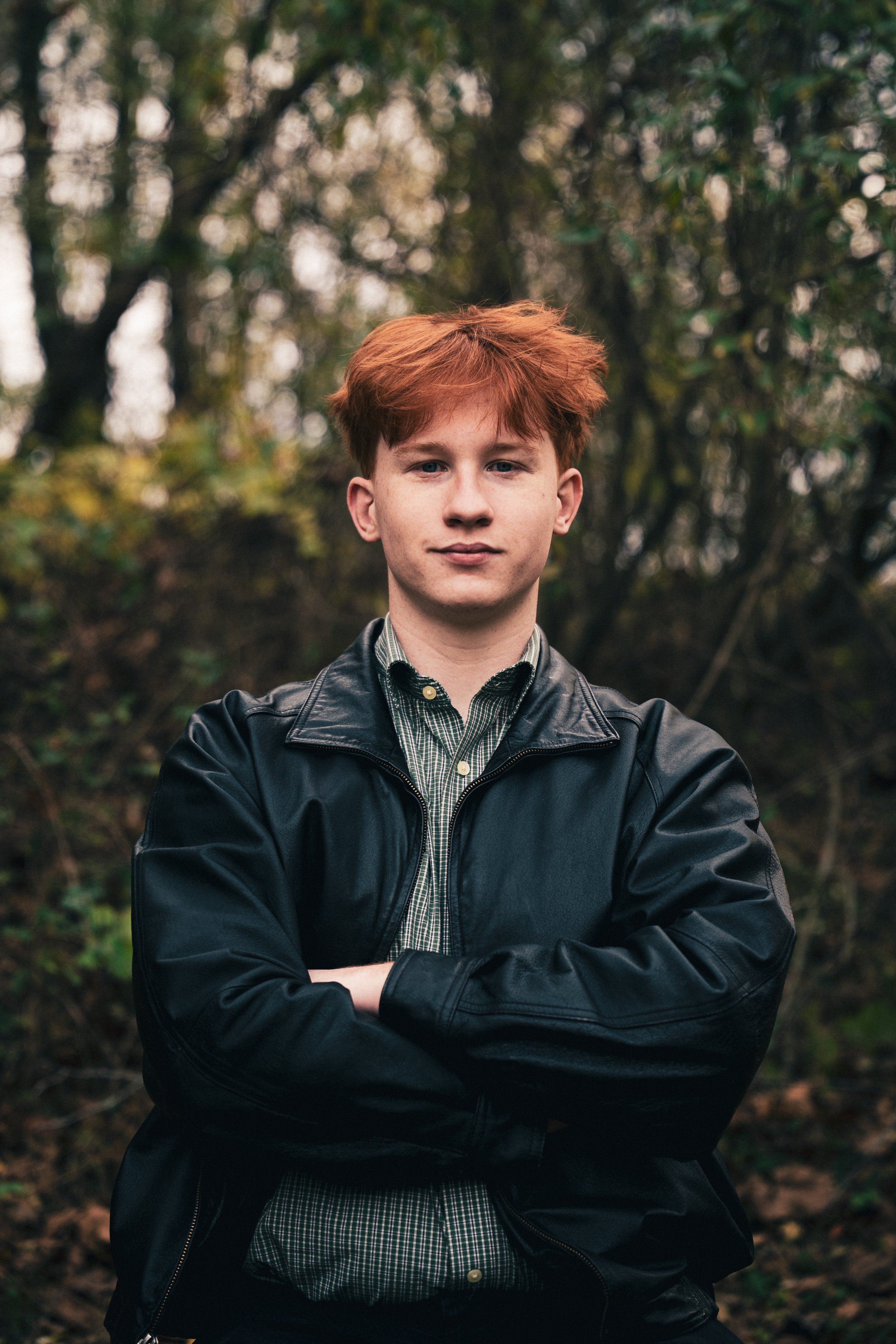 A young man with red hair and fair skin standing outdoors with arms crossed, wearing a black leather jacket and green checkered shirt, in a wooded area.