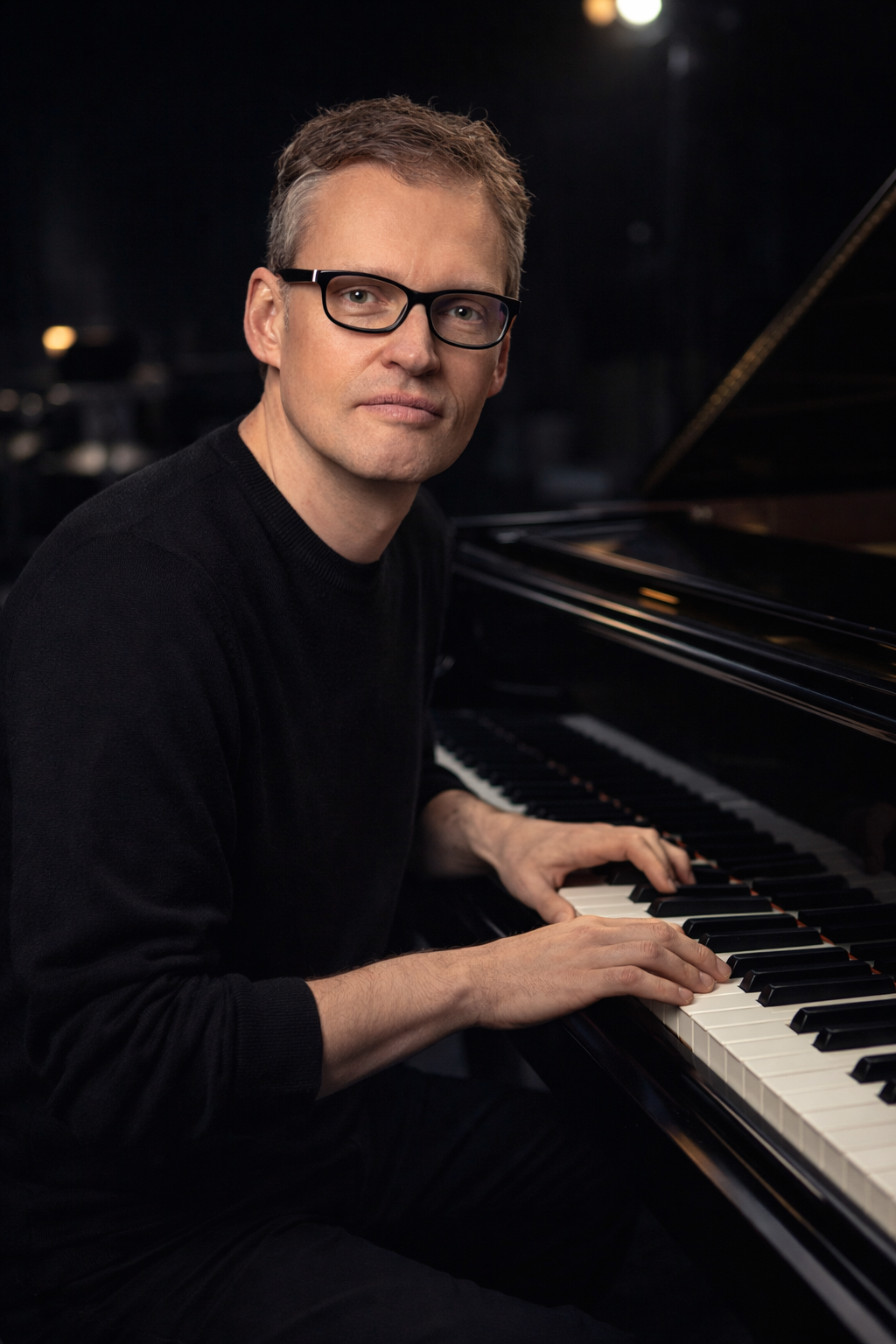 Mann spielt Klavier in dunklem Raum, trägt schwarze Kleidung und trägt Brille.