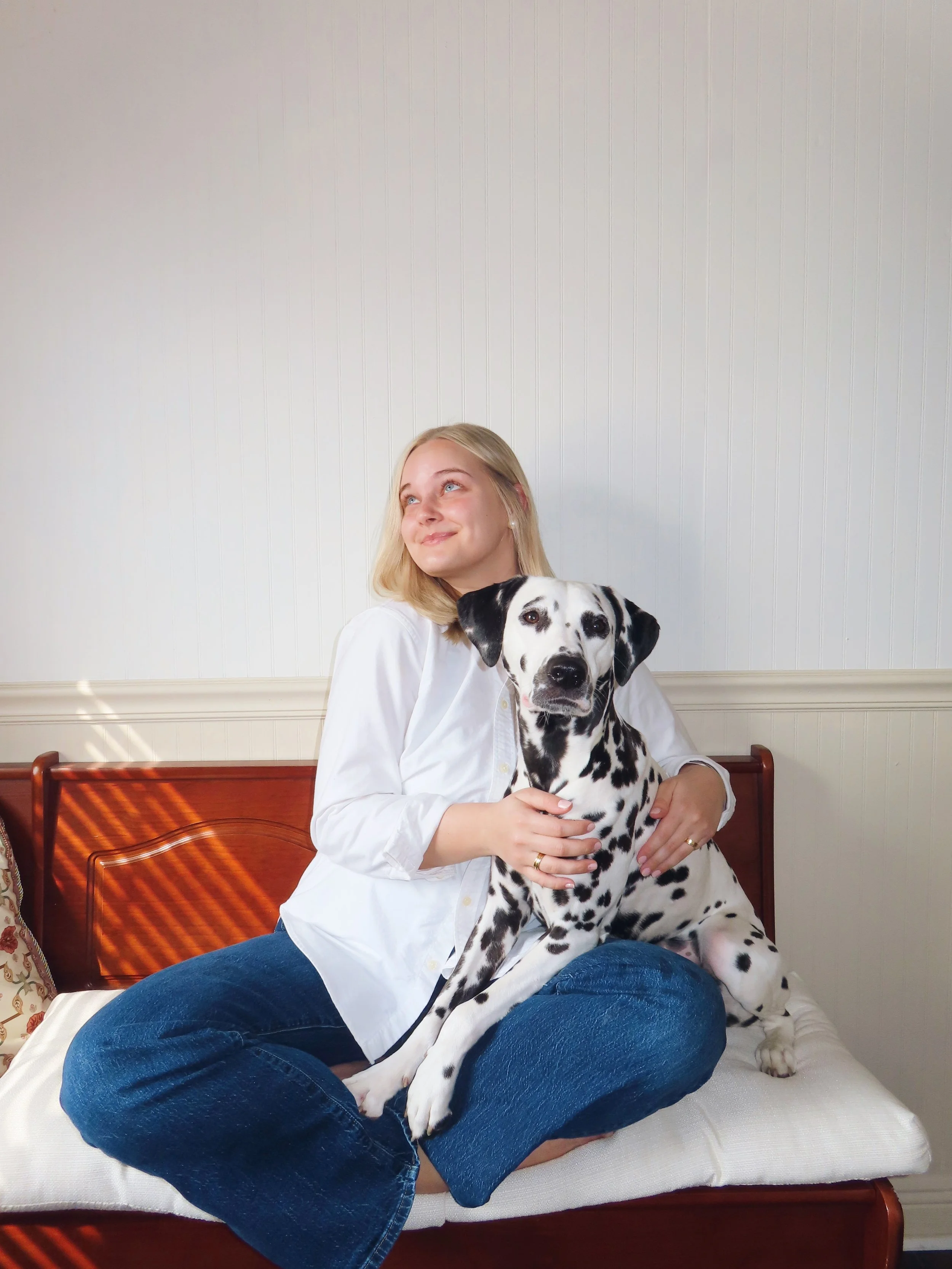 A woman sitting on a bench with a Dalmatian dog on her lap, indoors with white paneled walls.
