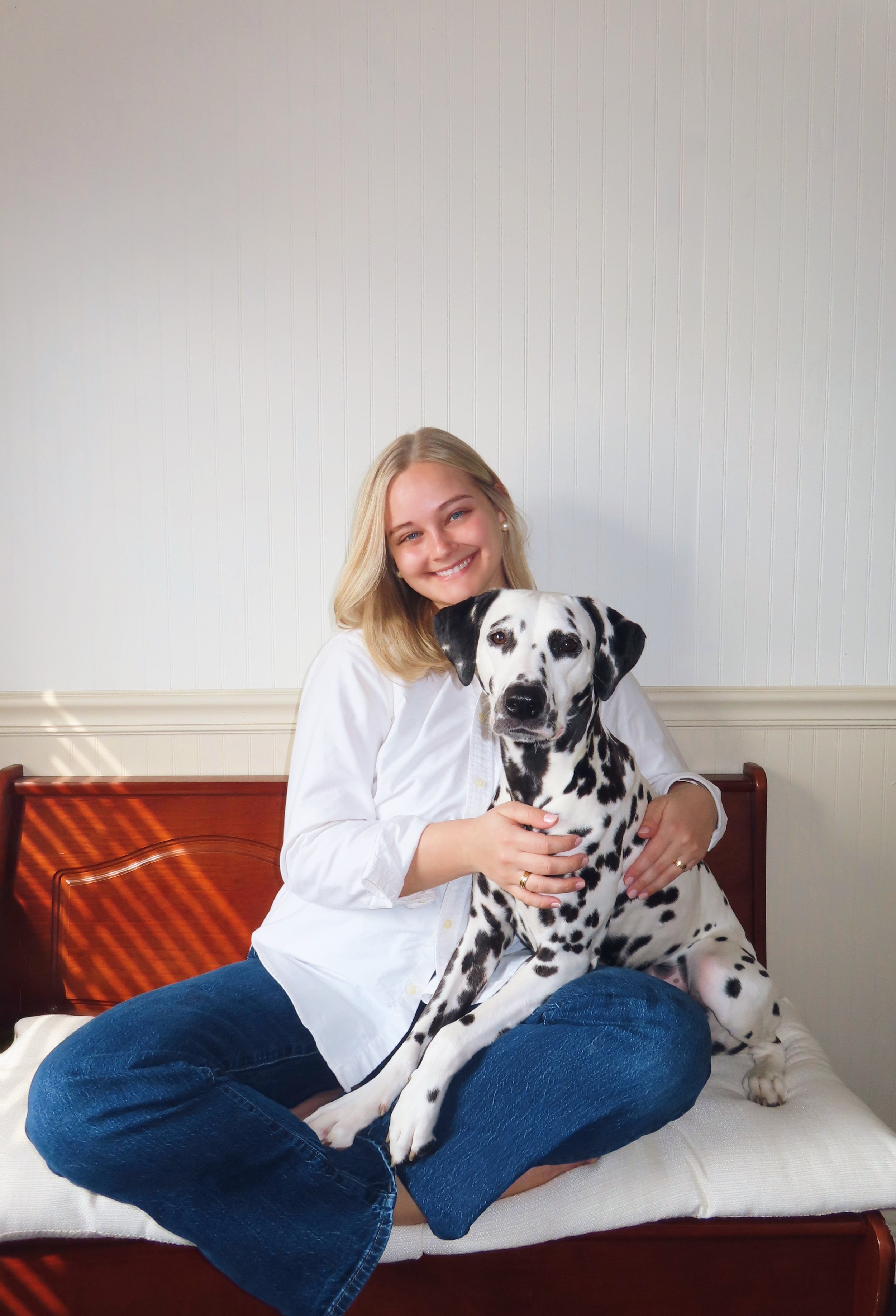 A smiling woman with blonde hair sitting cross-legged on a bench, hugging a Dalmatian dog, in front of a light-colored wall.