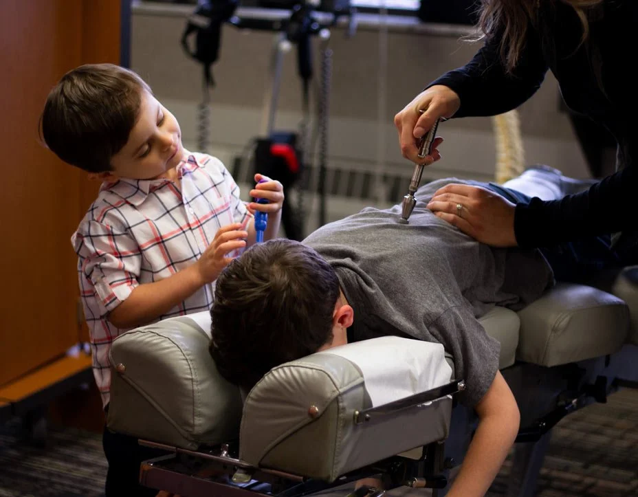 A chiropractor performs an adjustment on a man lying face down on a chiropractic table, while a young boy in a checkered shirt observes and holds a stethoscope in a professional medical setting.