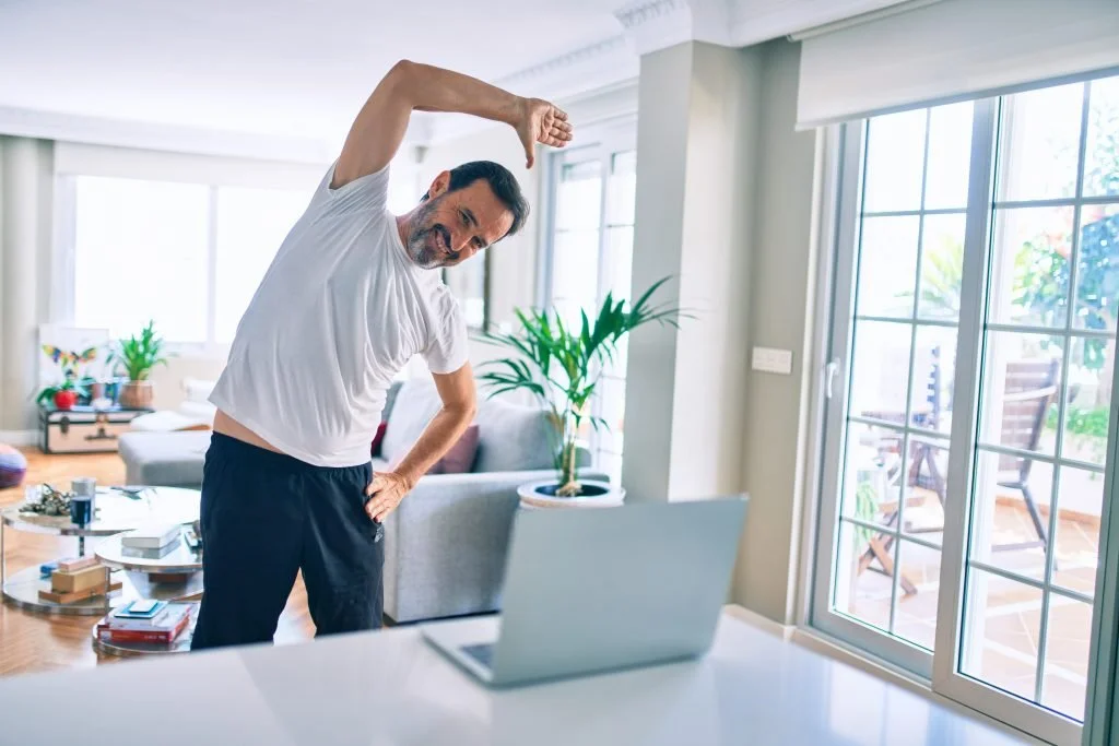 A man in a white T-shirt and black pants stretching with one arm over his head while standing in a bright living room near a laptop.