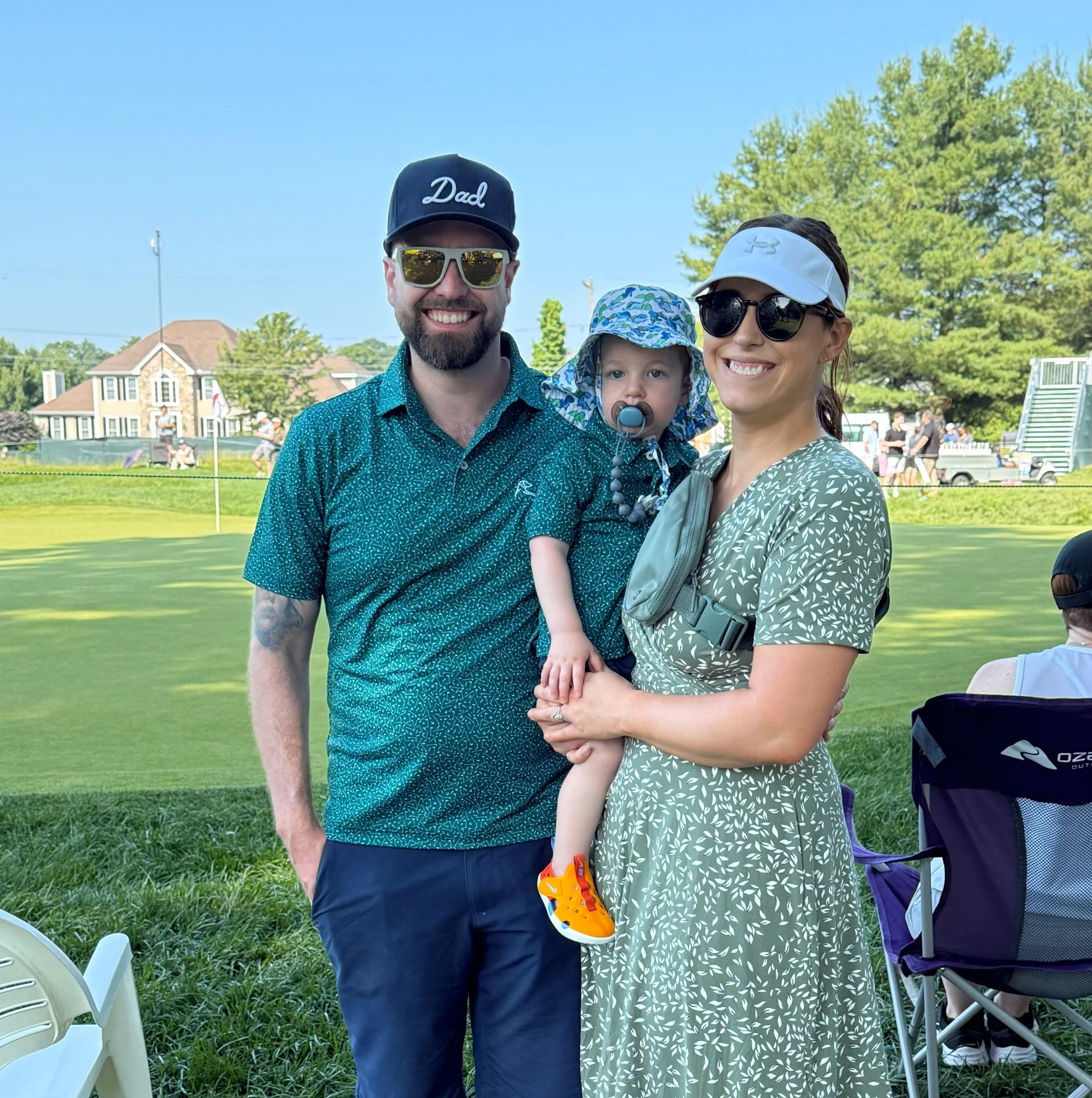 A happy family of three, with a man, woman, and young child, standing outdoors on a golf course on a sunny day. The man wears a cap that says 'Dad' and sunglasses. The woman wears a visor and sunglasses, and holds the child who has a pacifier and colorful hat. They are smiling at the camera.