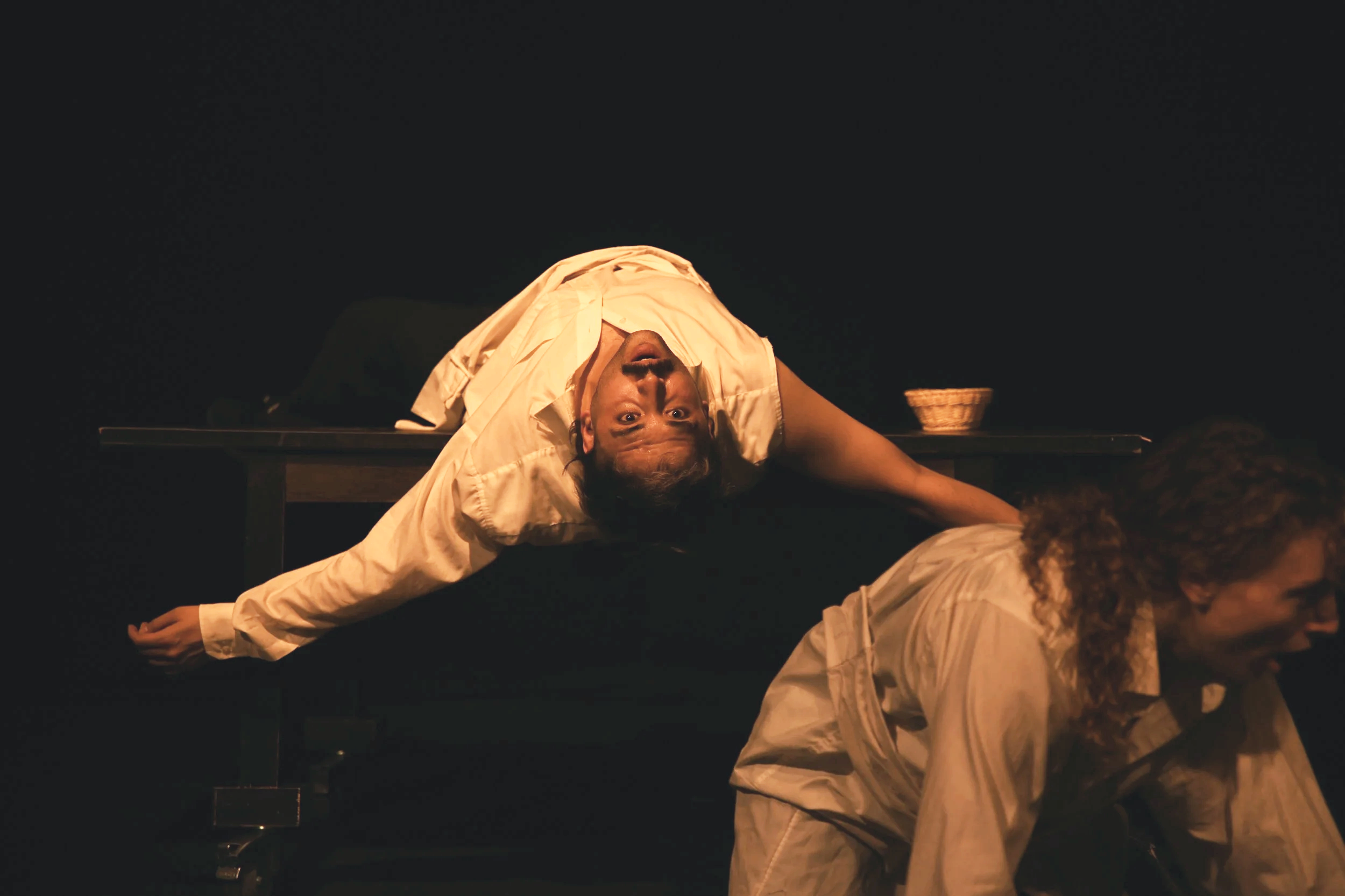 Two performers on stage, one lying horizontally on a table with her head hanging downward and looking towards the camera, the other kneeling on the ground in front of her, both dressed in light-colored clothing, with a dark background and a small bas