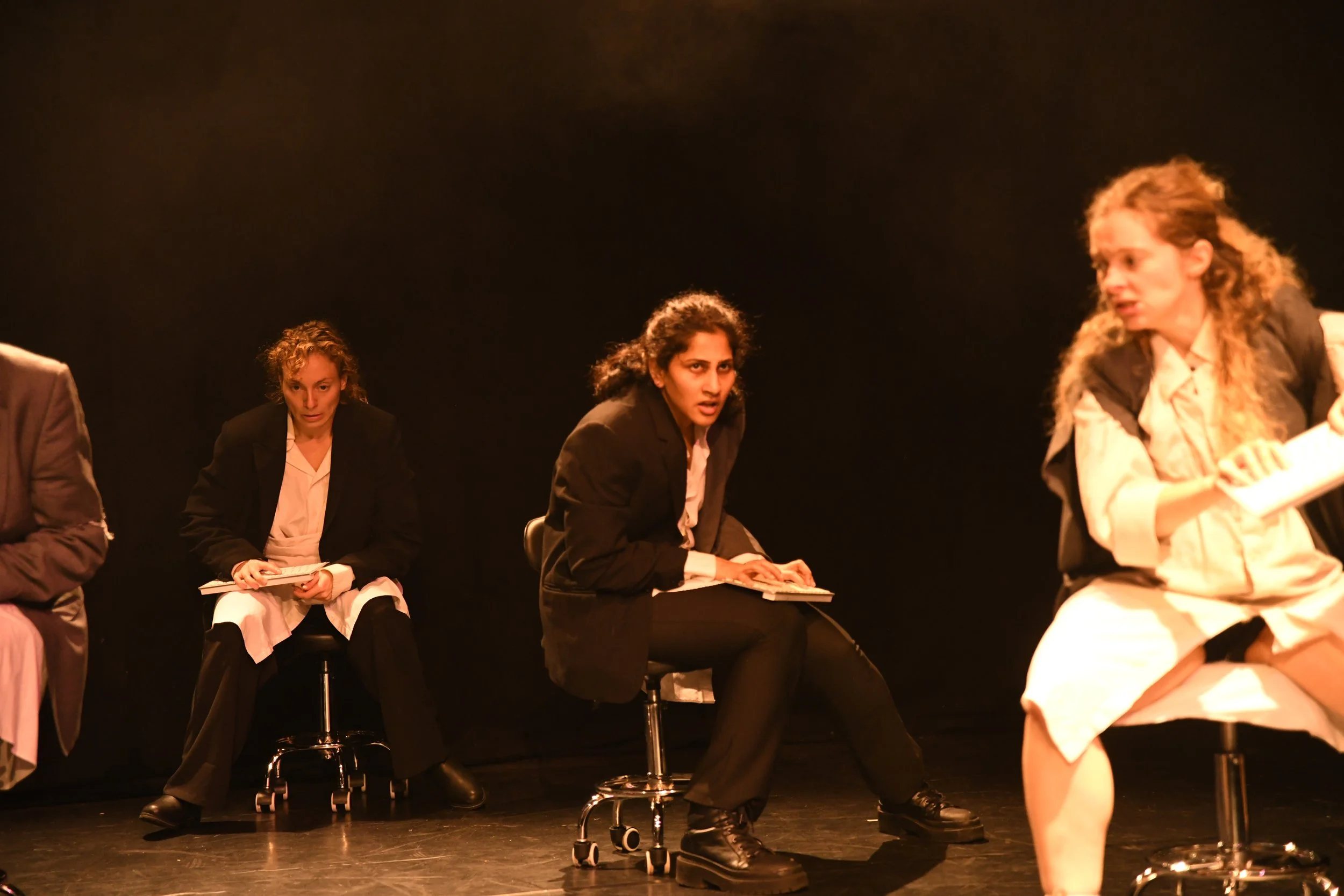 Four women in dark suits sitting on stools on a stage, appearing to be in a theatrical performance, with a dark background.