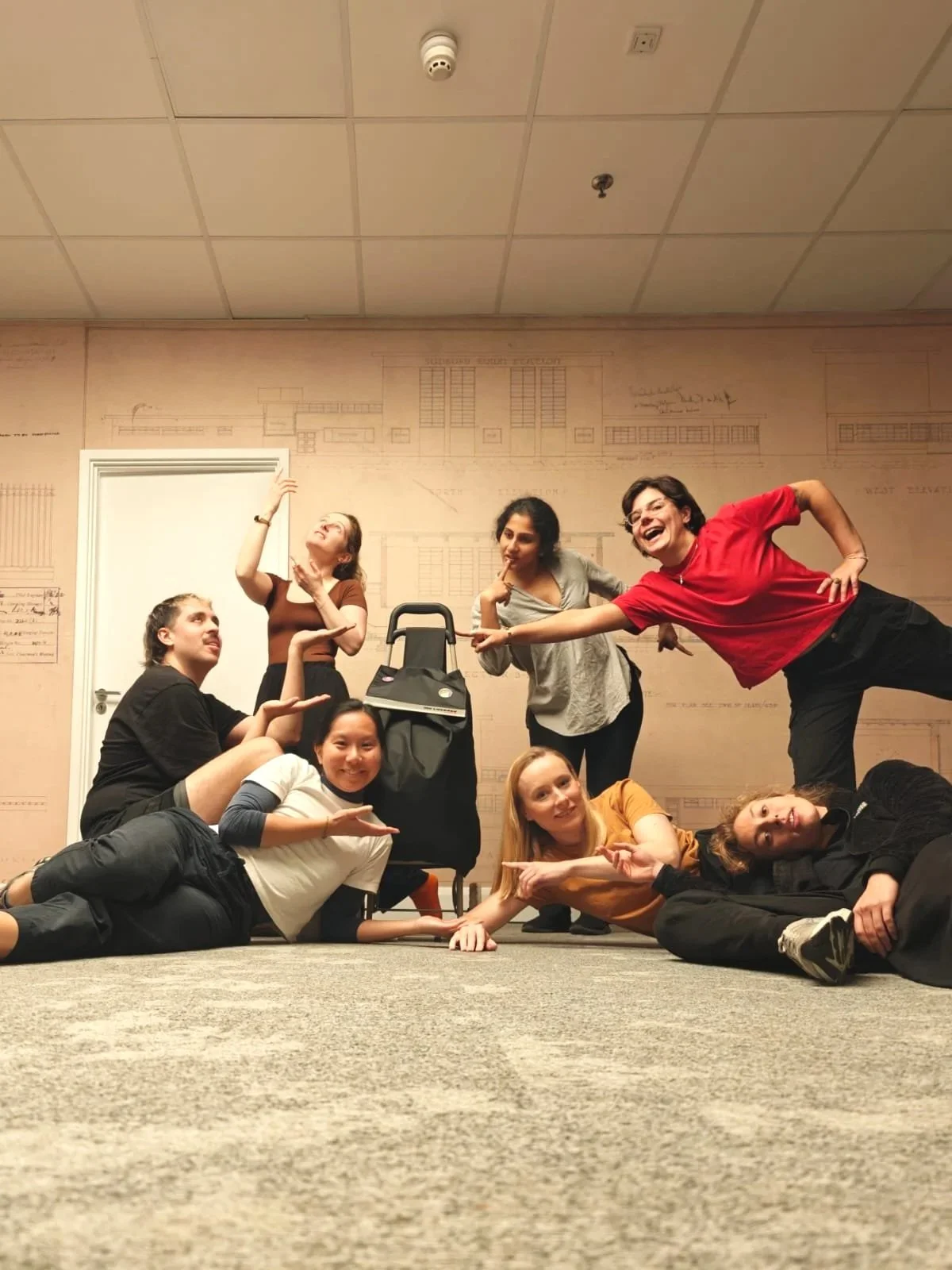Group of eight people posing playfully indoors against a beige wall with architectural drawings, some sitting or lying on the carpeted floor, one standing with hand on hip, one standing near a stroller, and three people making animated gestures.