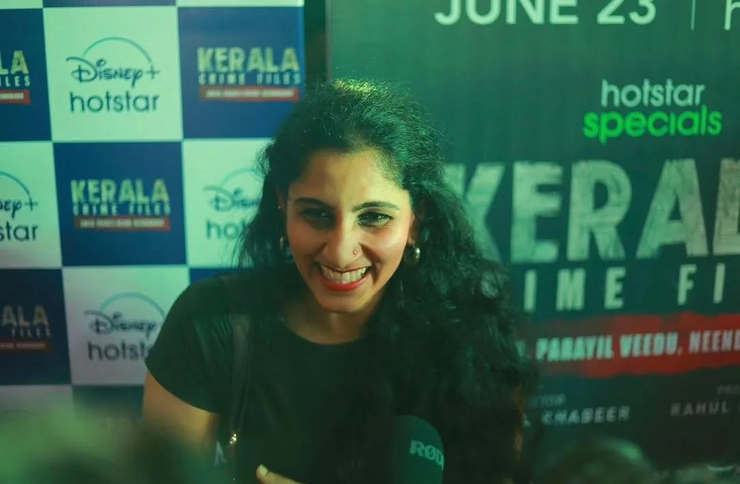 A woman with long curly black hair and a nose piercing smiling at the camera, holding a microphone, standing in front of a promotional backdrop for Kerala movie time. The backdrop includes logos for Disney+ Hotstar and Kerala Film Festival, with event details and dates.