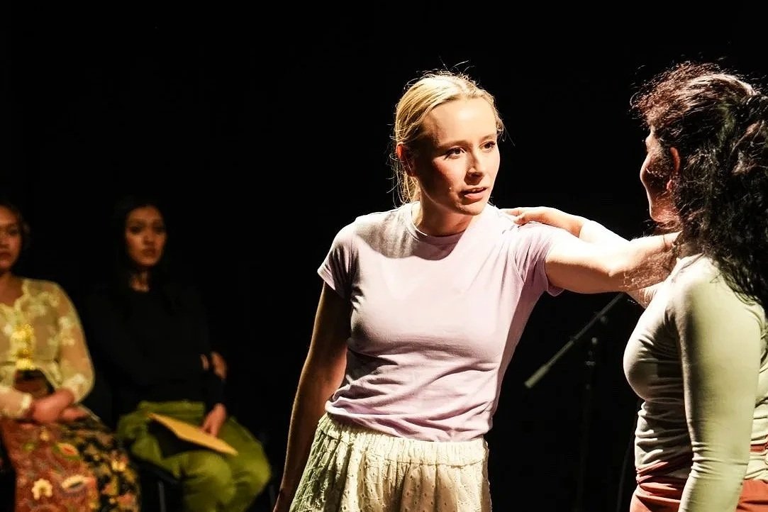 Two women engaged in an intense conversation during a theatrical performance, with three other women sitting in the background.