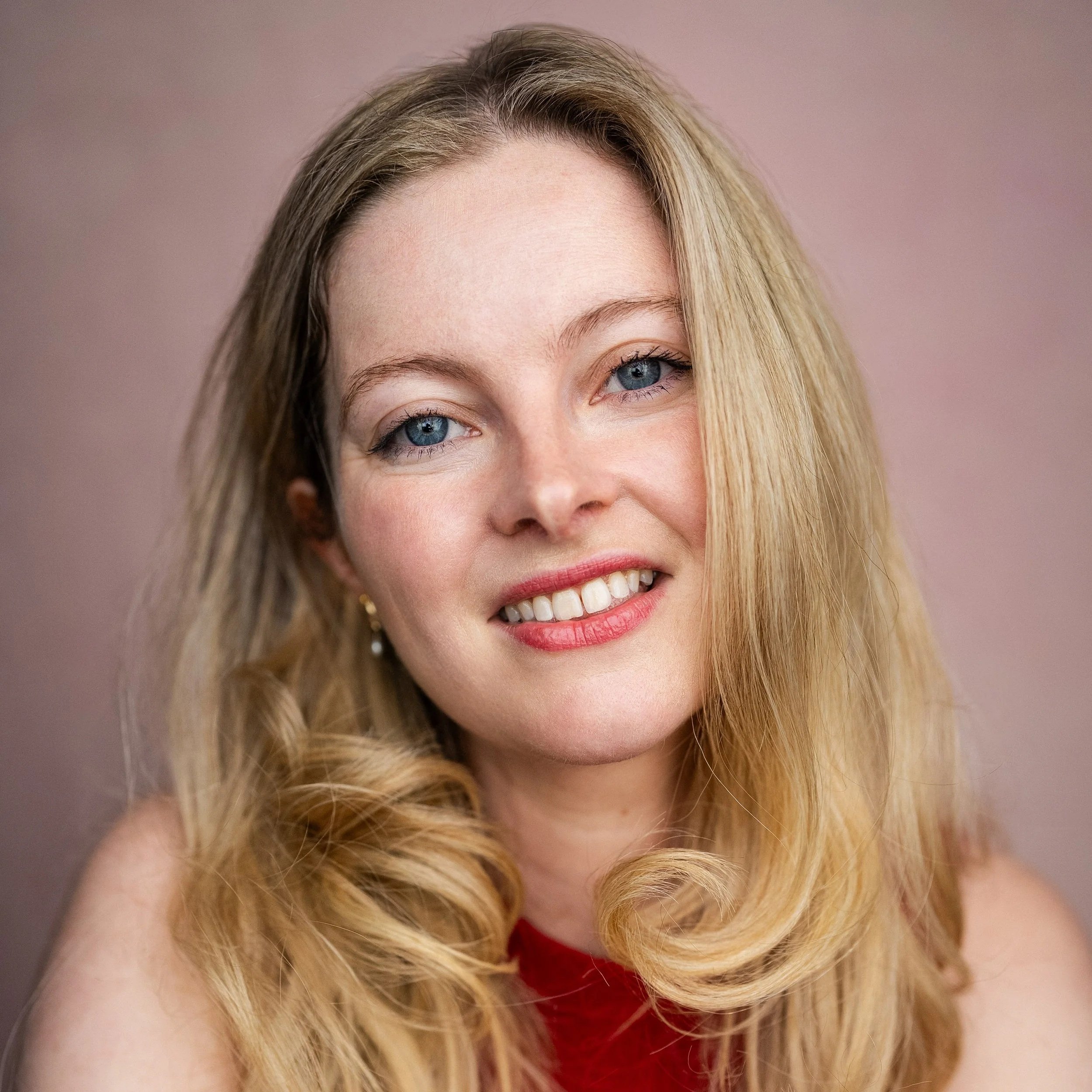 Close-up of a smiling woman with wavy blonde hair, blue eyes, and fair skin in front of a soft pink background.