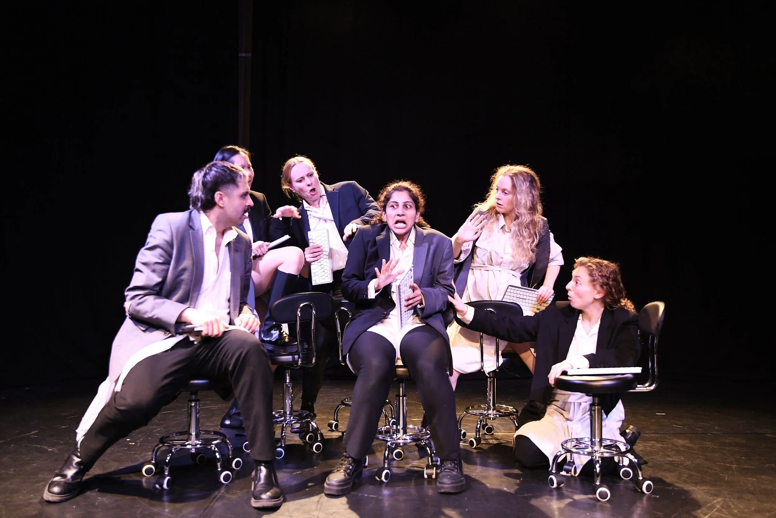 Six actors in business attire participating in a theatrical performance on stage, with a black background.