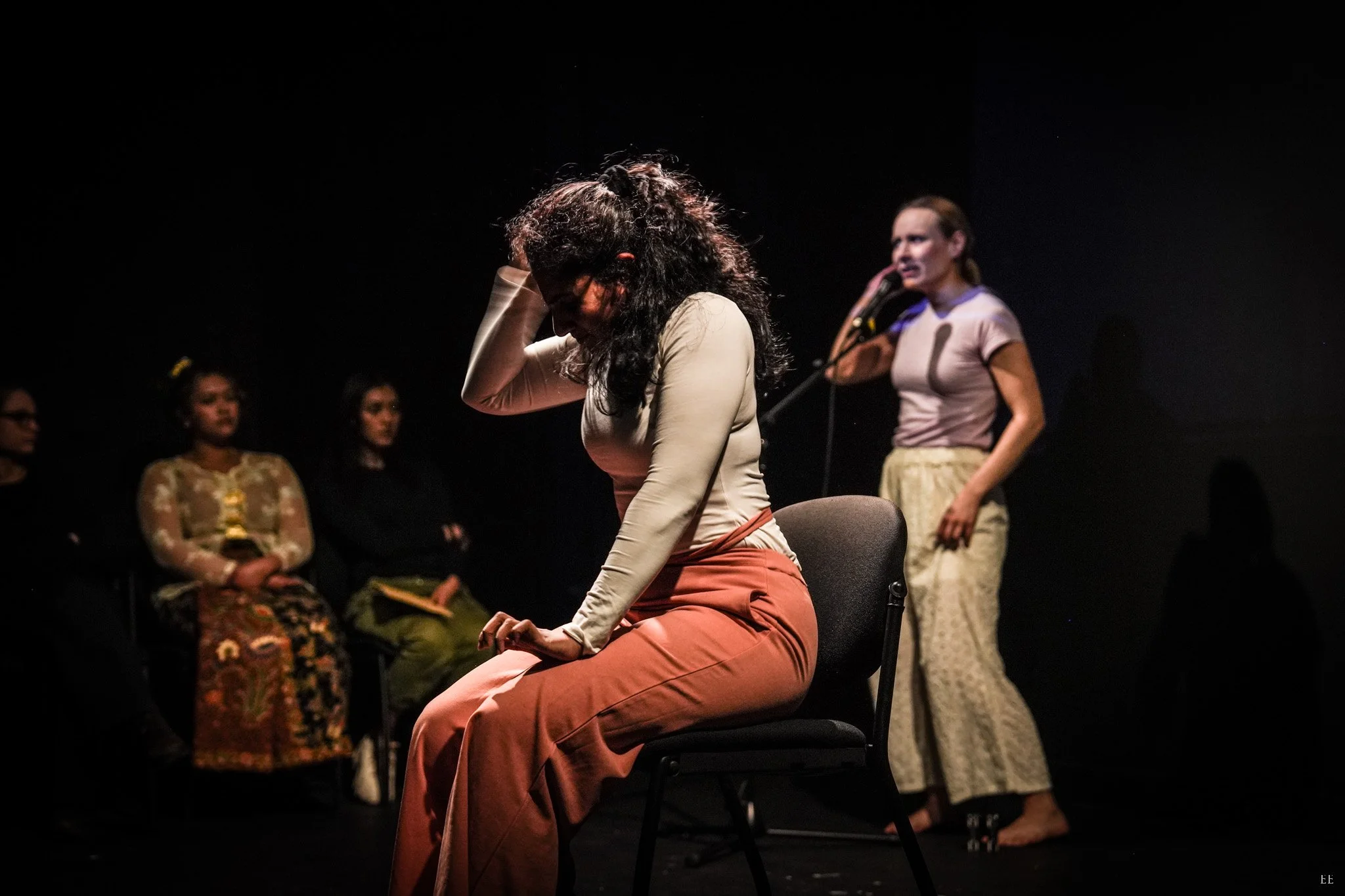 A woman with curly hair sitting on a chair, holding her head with one hand, in a theatrical setting with a spotlight, other women sitting in the background and one woman standing and speaking into a microphone.