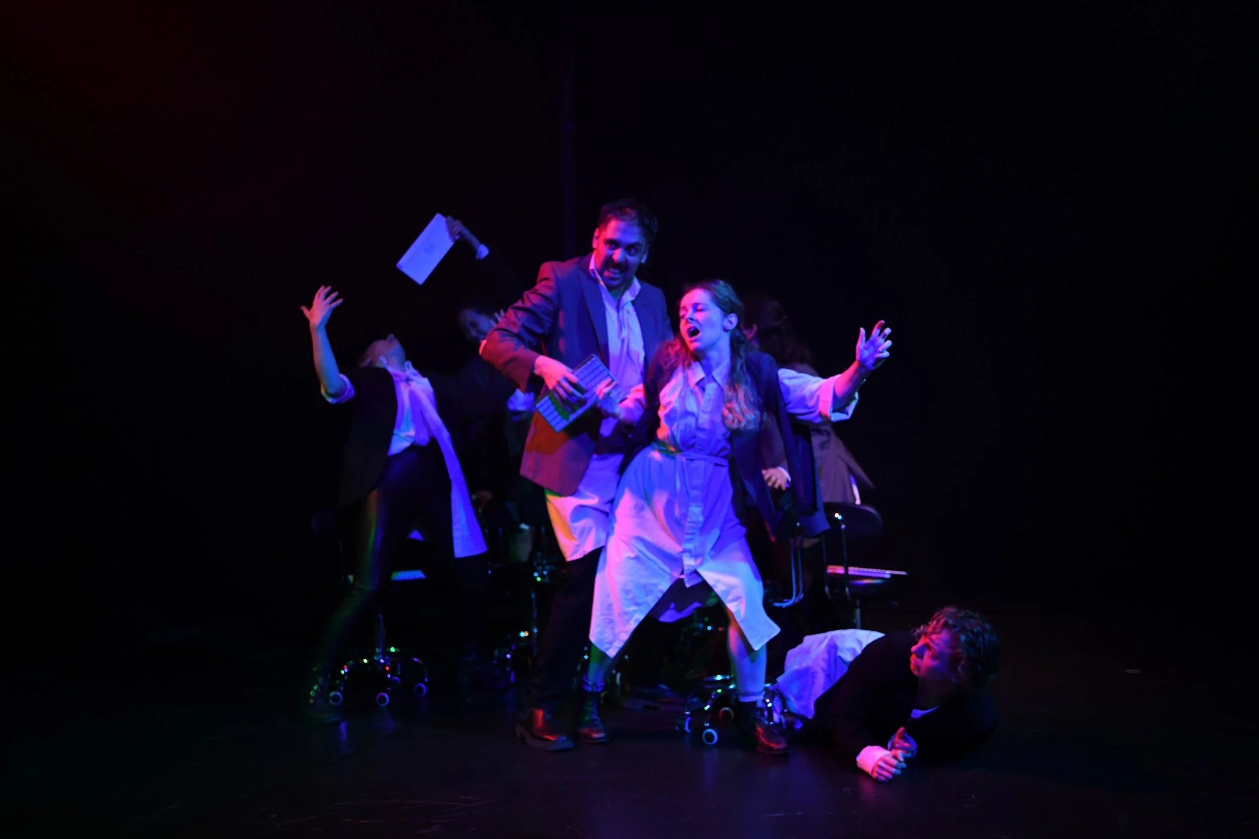 People on stage performing in a theatrical scene under colorful lighting, with one person lying on the floor and others expressing dramatic emotions.