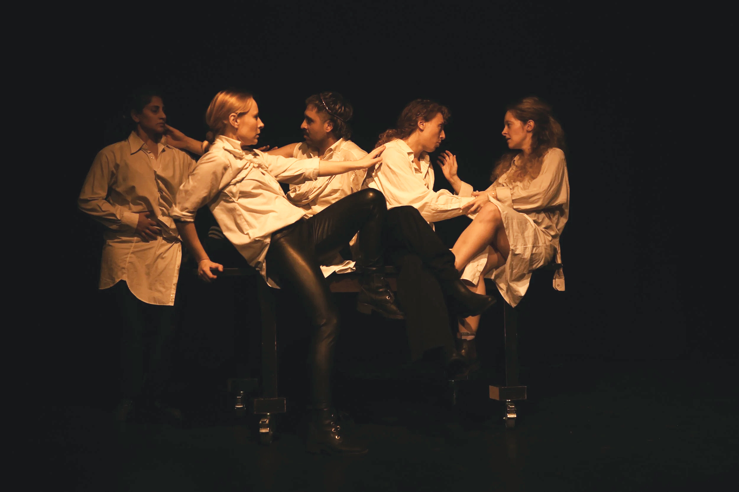 Six women on stage, five sitting on a bench and one standing, engaged in a theatrical performance with intense expressions, in dim lighting.