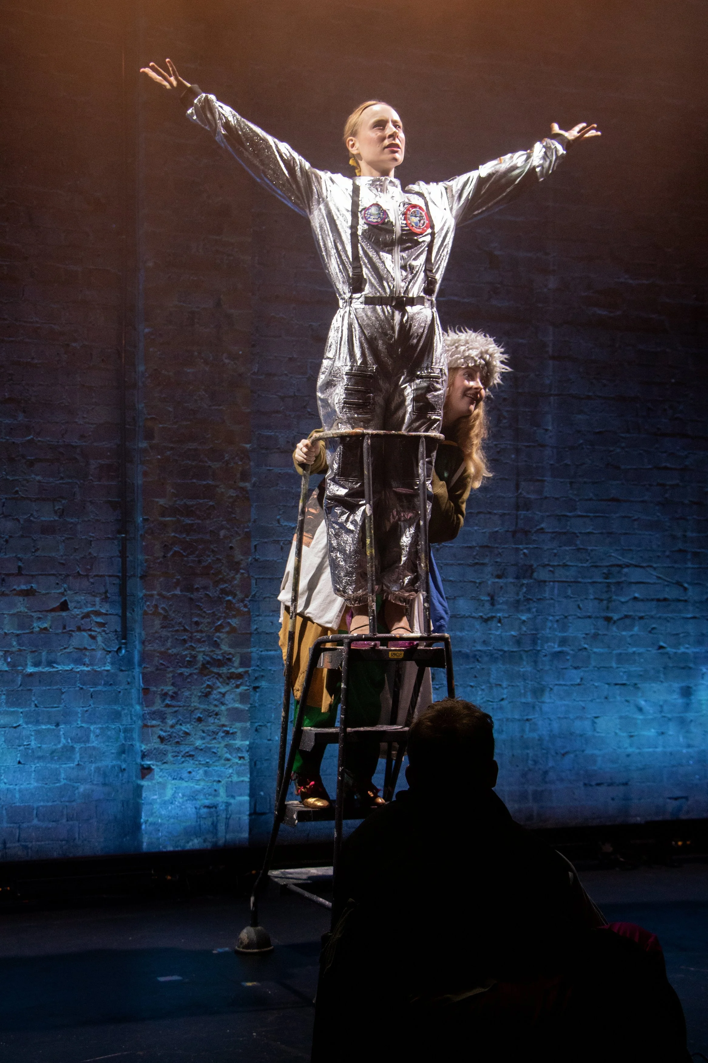 A woman in a silver jumpsuit standing on a platform with arms outstretched, supported by another woman in costume, with a blue brick wall in the background.