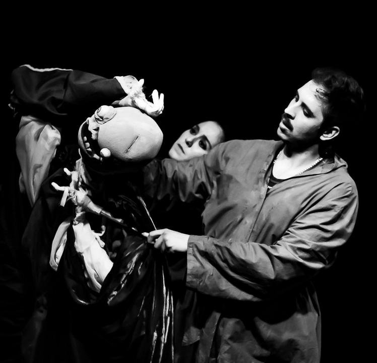 Two young adults perform a puppet show with a hand puppet resembling a face, using dark costumes and a black background.