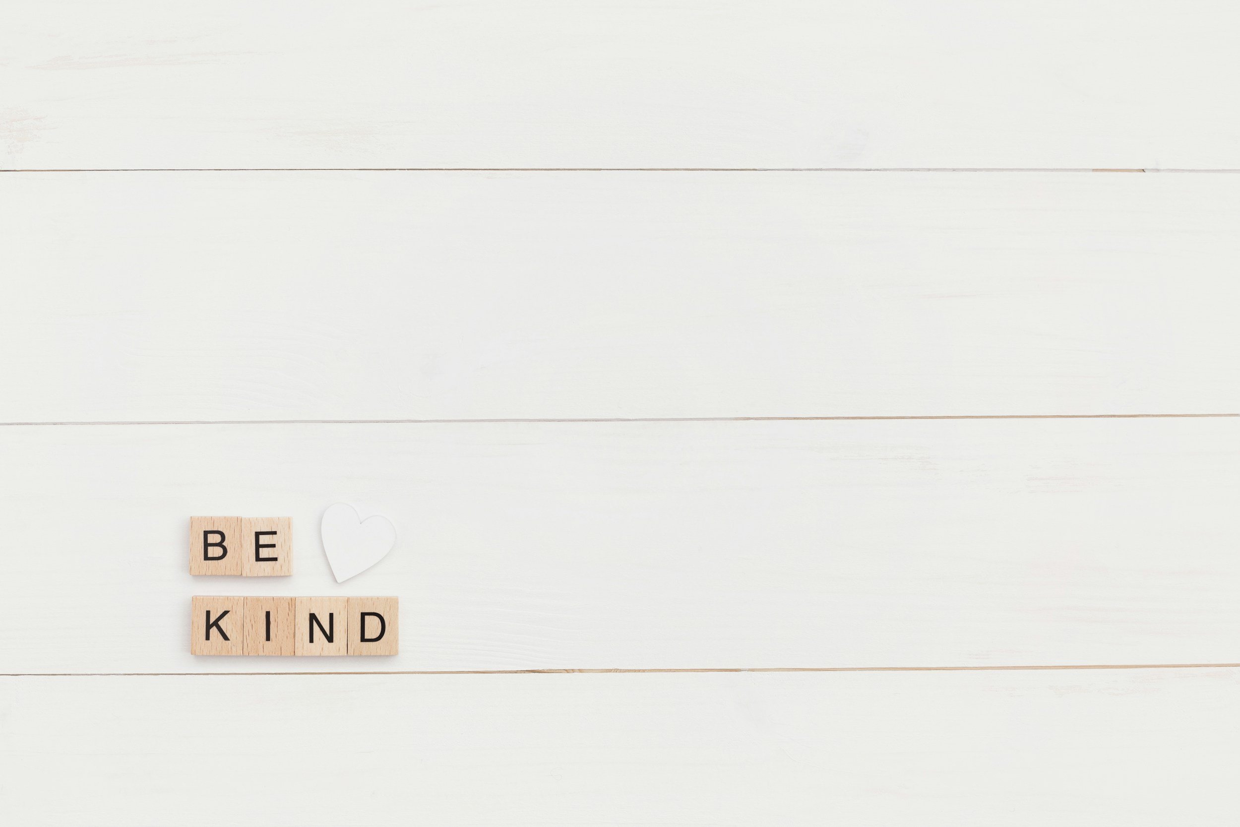 Tarjetas con las palabras 'BE' y 'KIND' en letras negras, acompañadas de un corazón blanco de papel sobre fondo de tablas blancas.