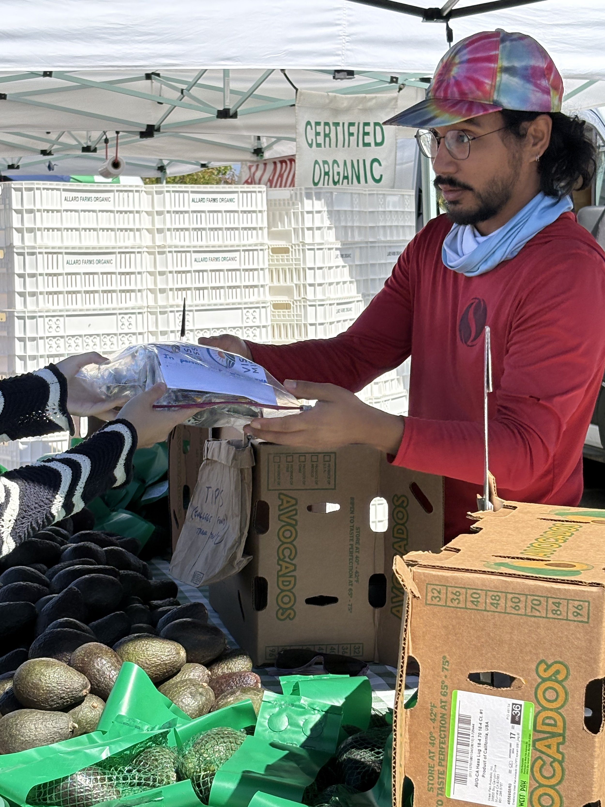A man wearing glasses, a colorful tie-dye cap, and a red shirt is handing a produce item to a customer at an outdoor market stall. The stall has a sign that reads 'Certified Organic,' and there are white crates with labels that say 'Allard Farms Orga