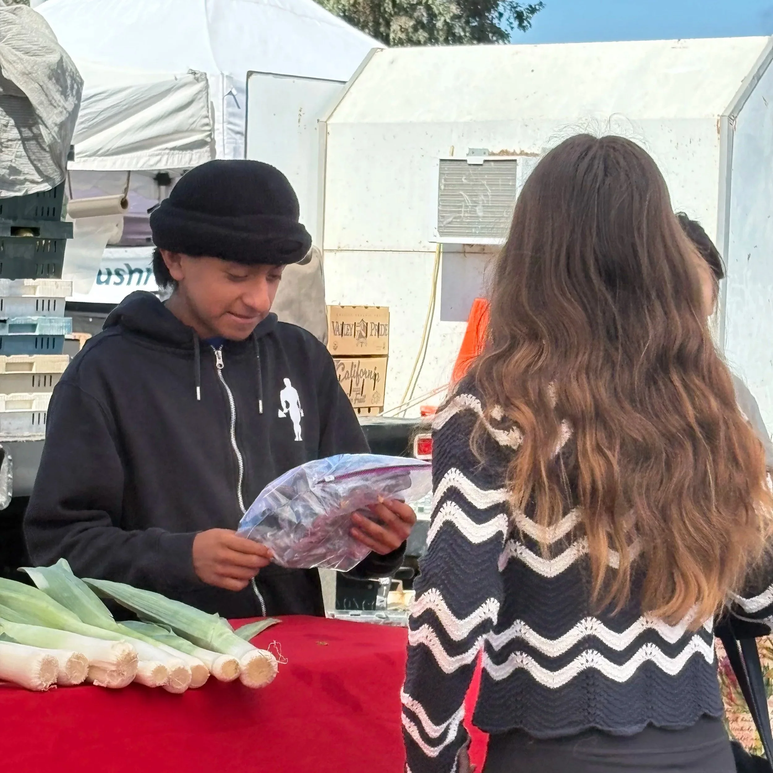A woman with long brown hair wearing a black and white chevron patterned sweater and an orange shirt is standing in front of a vendor stand at an outdoor market. The stand has a display of white vegetables, likely parsnips or similar, arranged on a r
