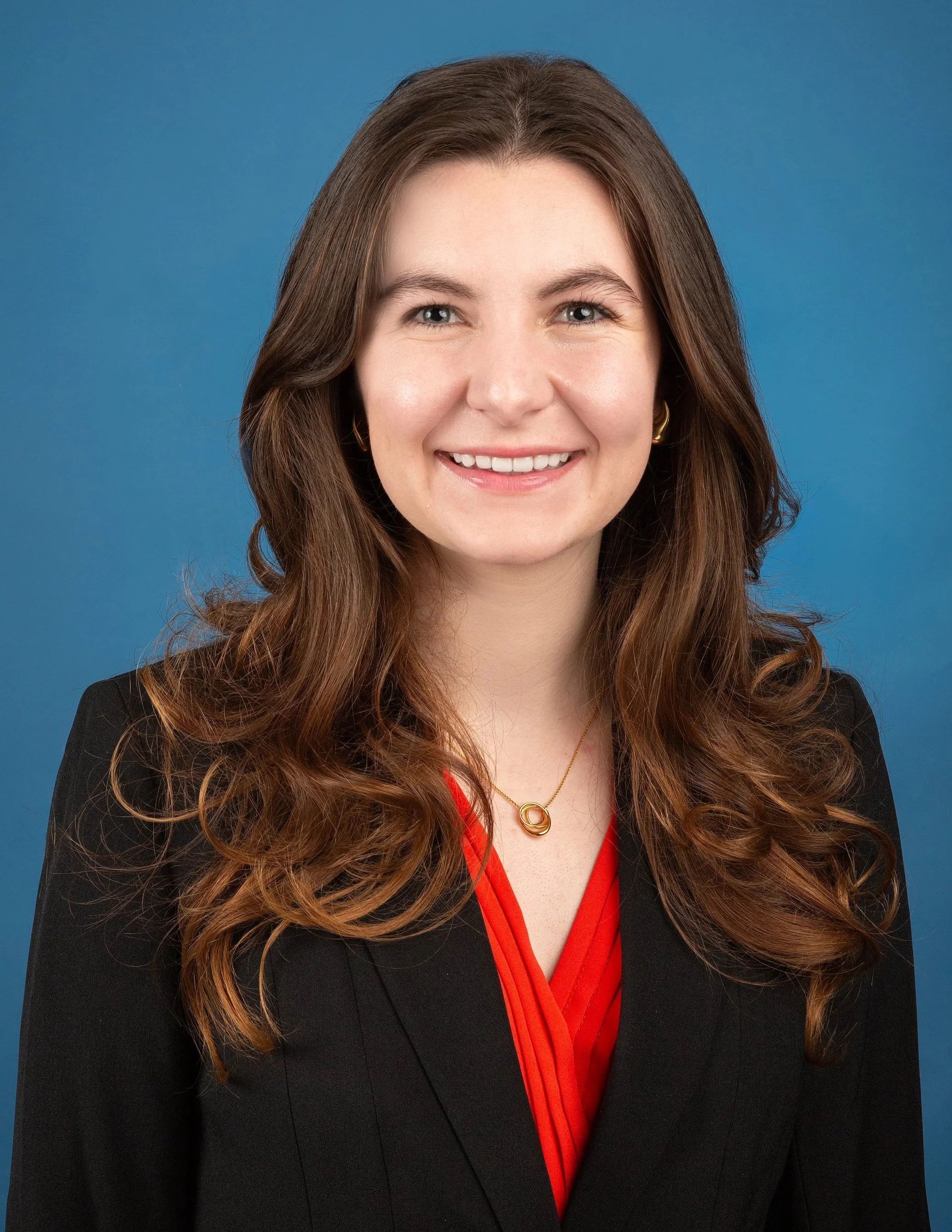 Close-up headshot of a young woman with long wavy brown hair, smiling, wearing a black blazer, a red blouse with a V-neck, and gold jewelry, against a solid blue background.