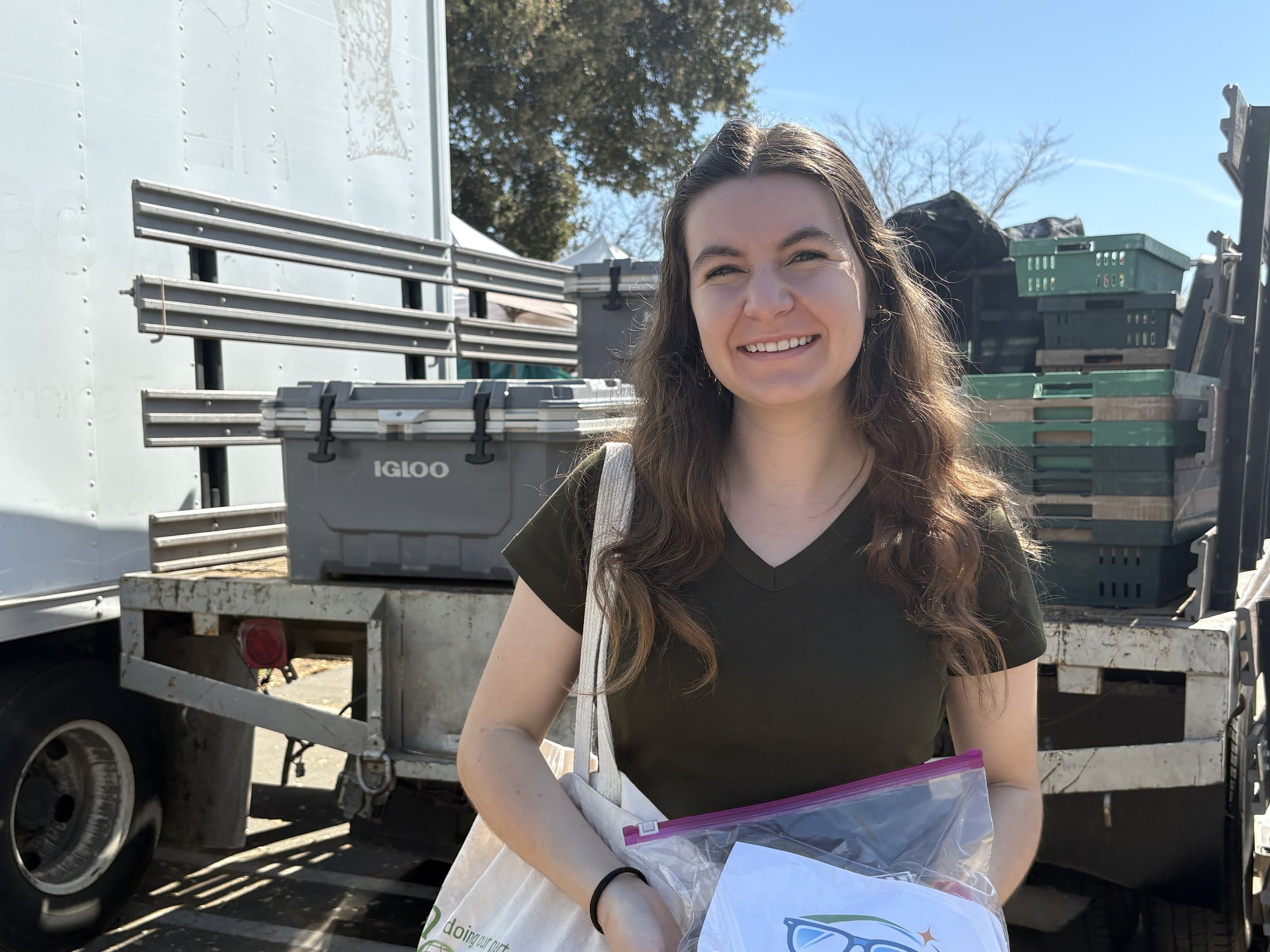 A smiling young woman with long curly hair holding a bag with a logo that says 'VISION MISSION' , standing outdoors near a trailer with stacked plastic crates and bins.