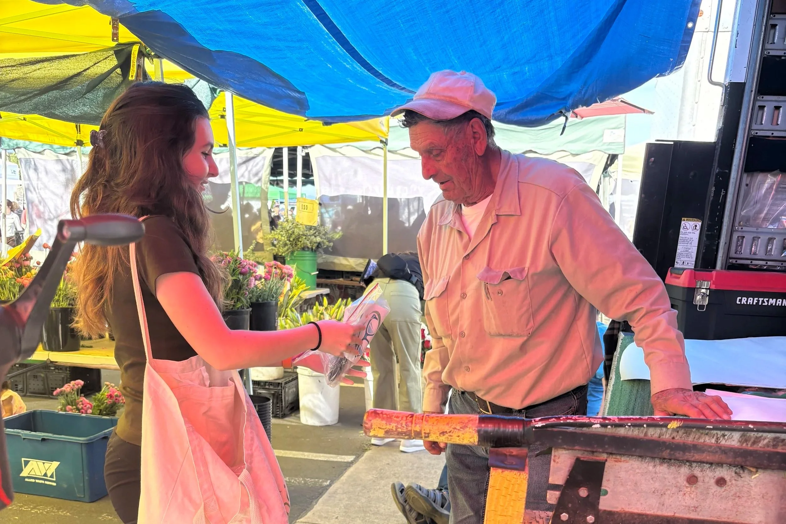 A young woman was giving money to an older man at a market. The man is wearing a pink cap and a pinkish shirt, while the woman has long brown hair and is wearing a dark-colored t-shirt. There are colorful tents in the background with various flowers 