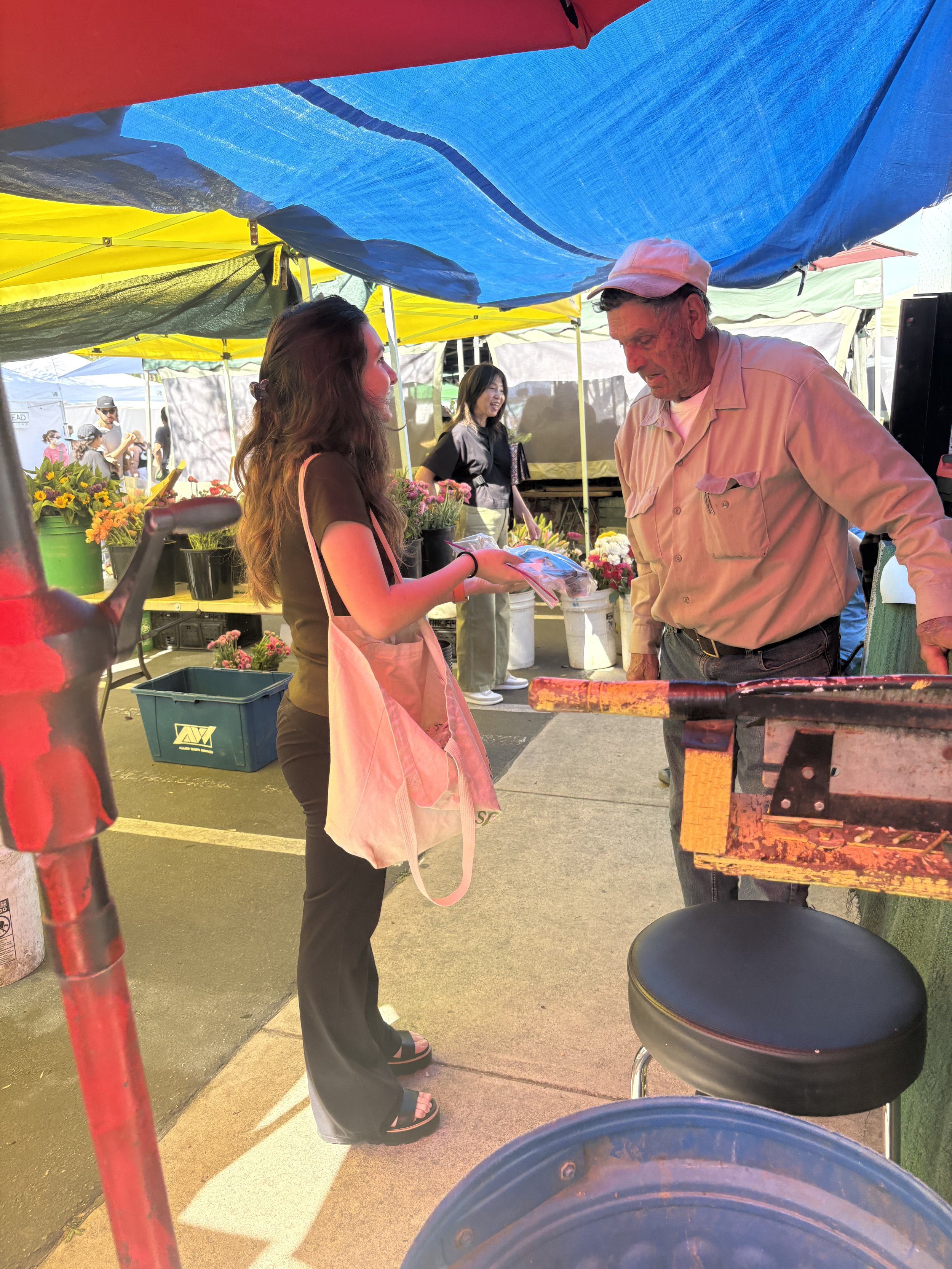 A young woman with long brown hair, wearing a black top and black pants, is handing a plastic bag to an older man with glasses and a cap at a flower market stall. The stall has colorful flowers and is covered with bright blue, yellow, and red tents. 