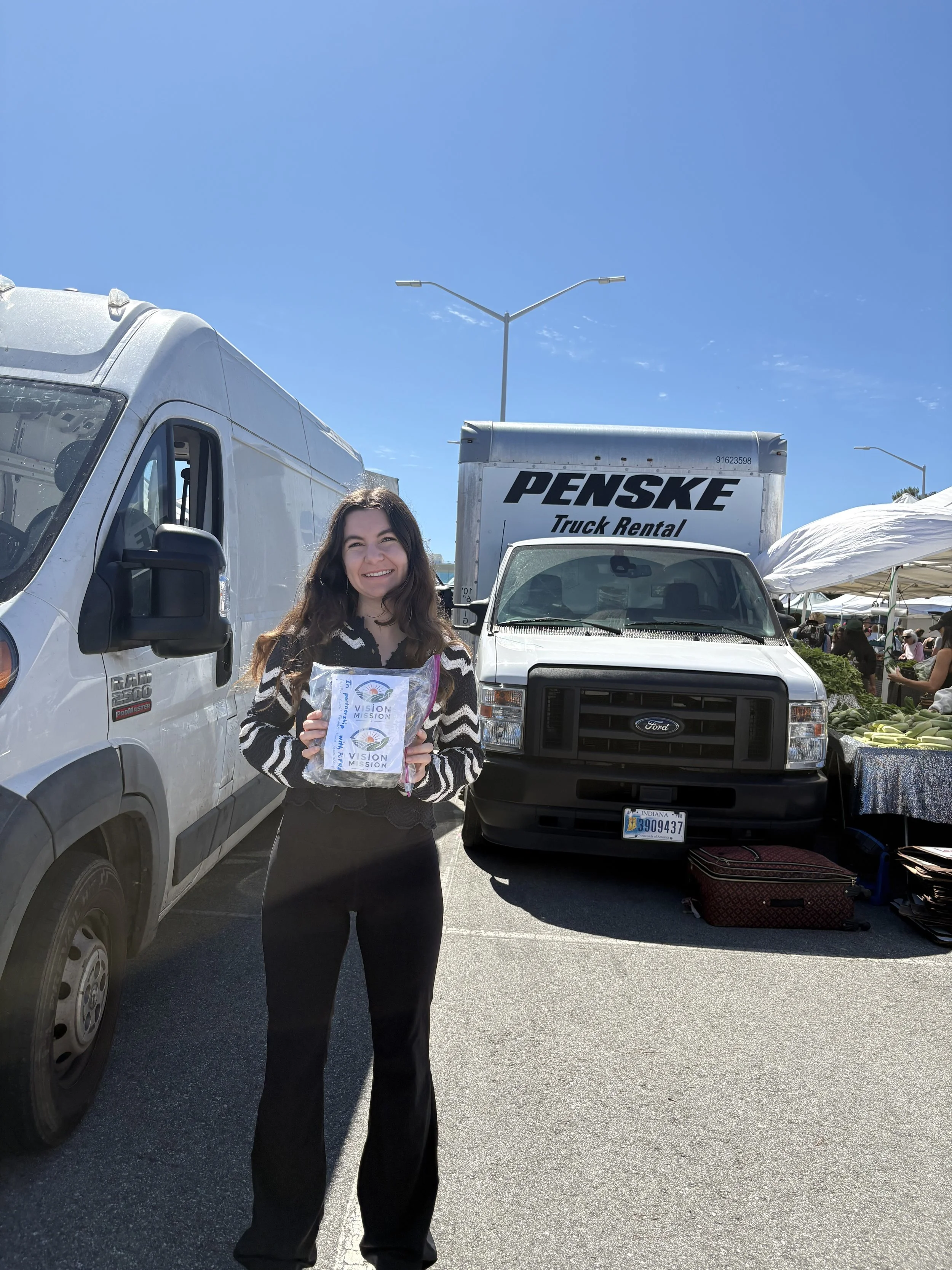 A smiling woman holding a plastic bag with a logo, standing in front of trucks and market tents at an outdoor market on a sunny day.