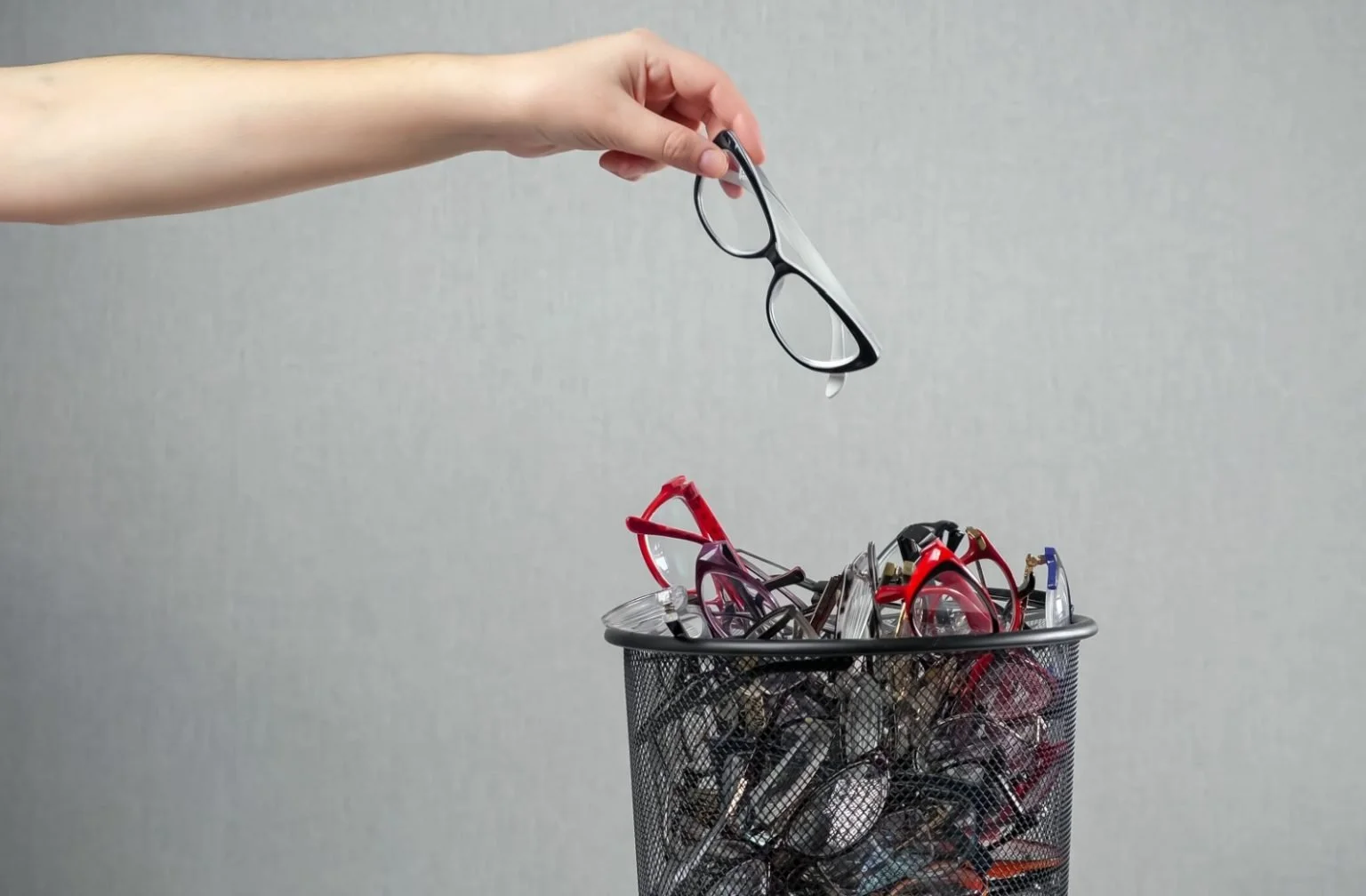 A hand holding a black and clear eyeglasses frame above a trash can filled with various discarded glasses and sunglasses.
