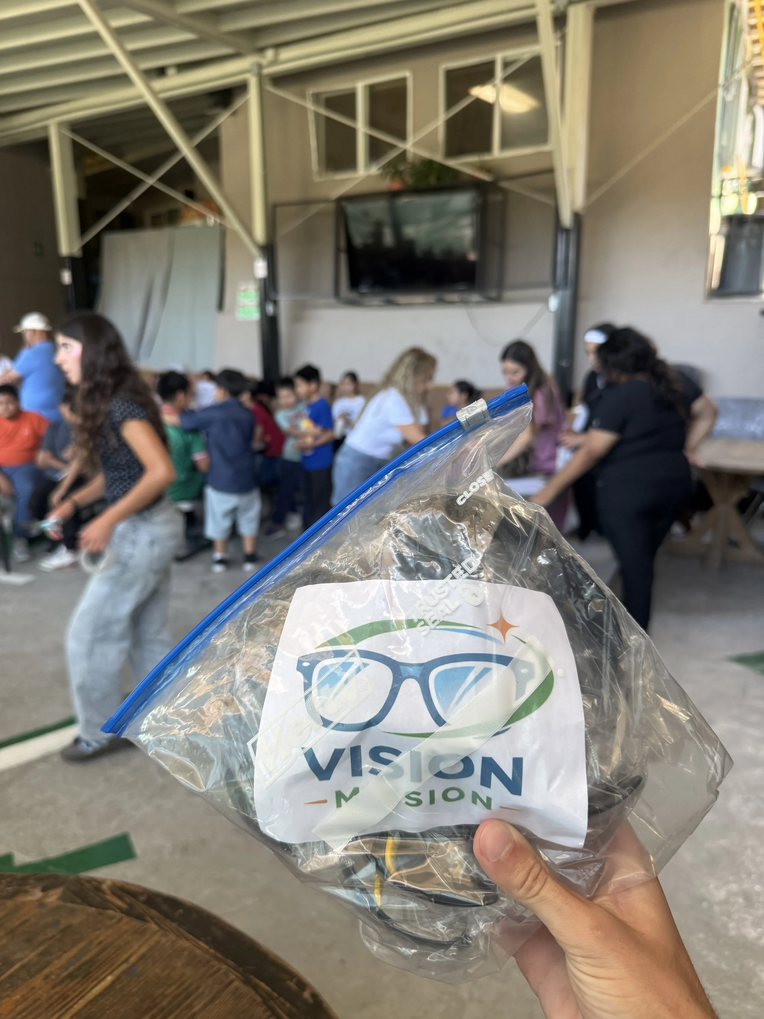 Hand holding a plastic bag with the words 'VISION' and 'MISION' in a crowded room with people, a tv, and a structure overhead.