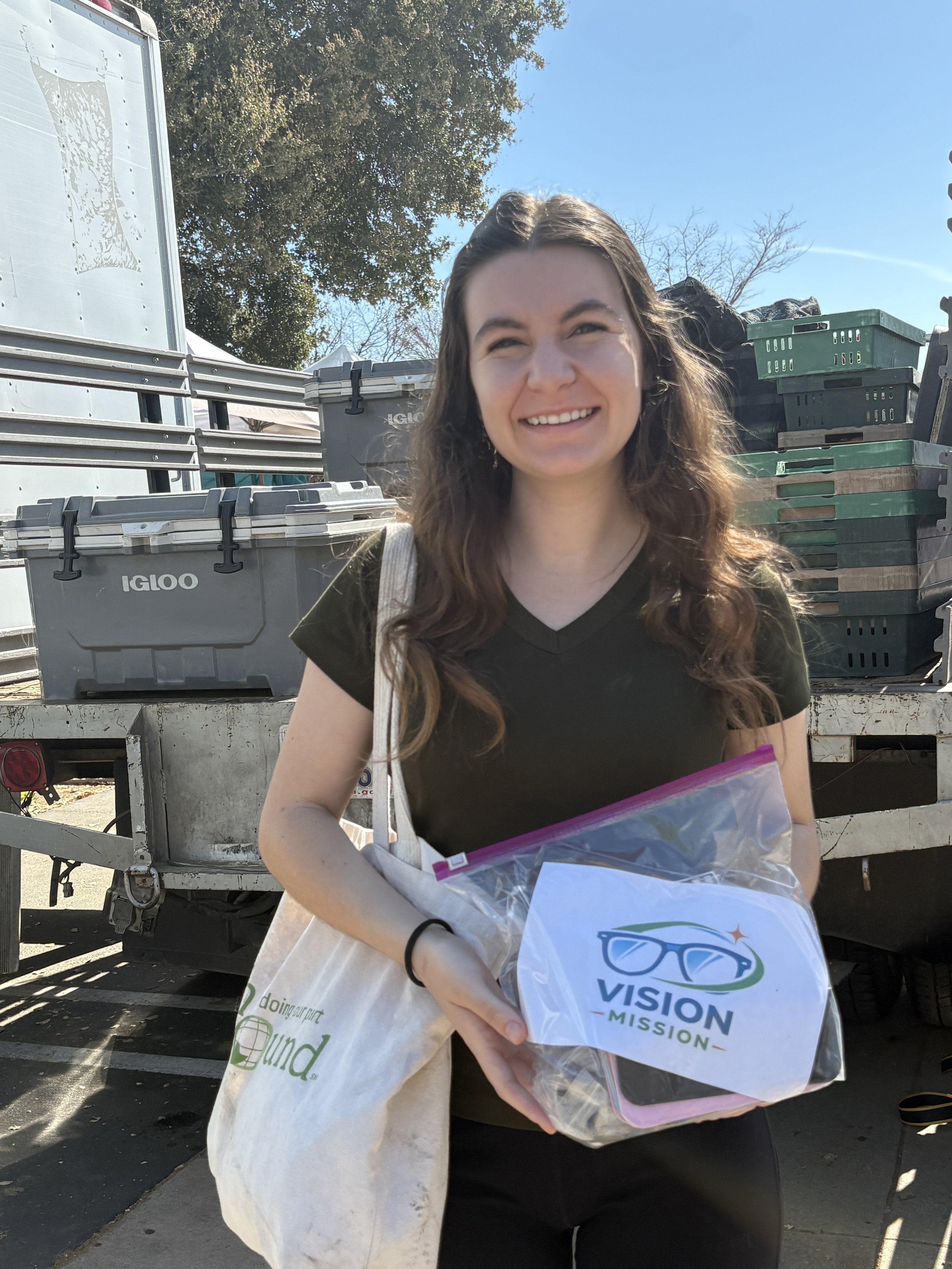 A smiling young woman with long curly hair holding a bag with a logo that says 'VISION MISSION' , standing outdoors near a trailer with stacked plastic crates and bins.
