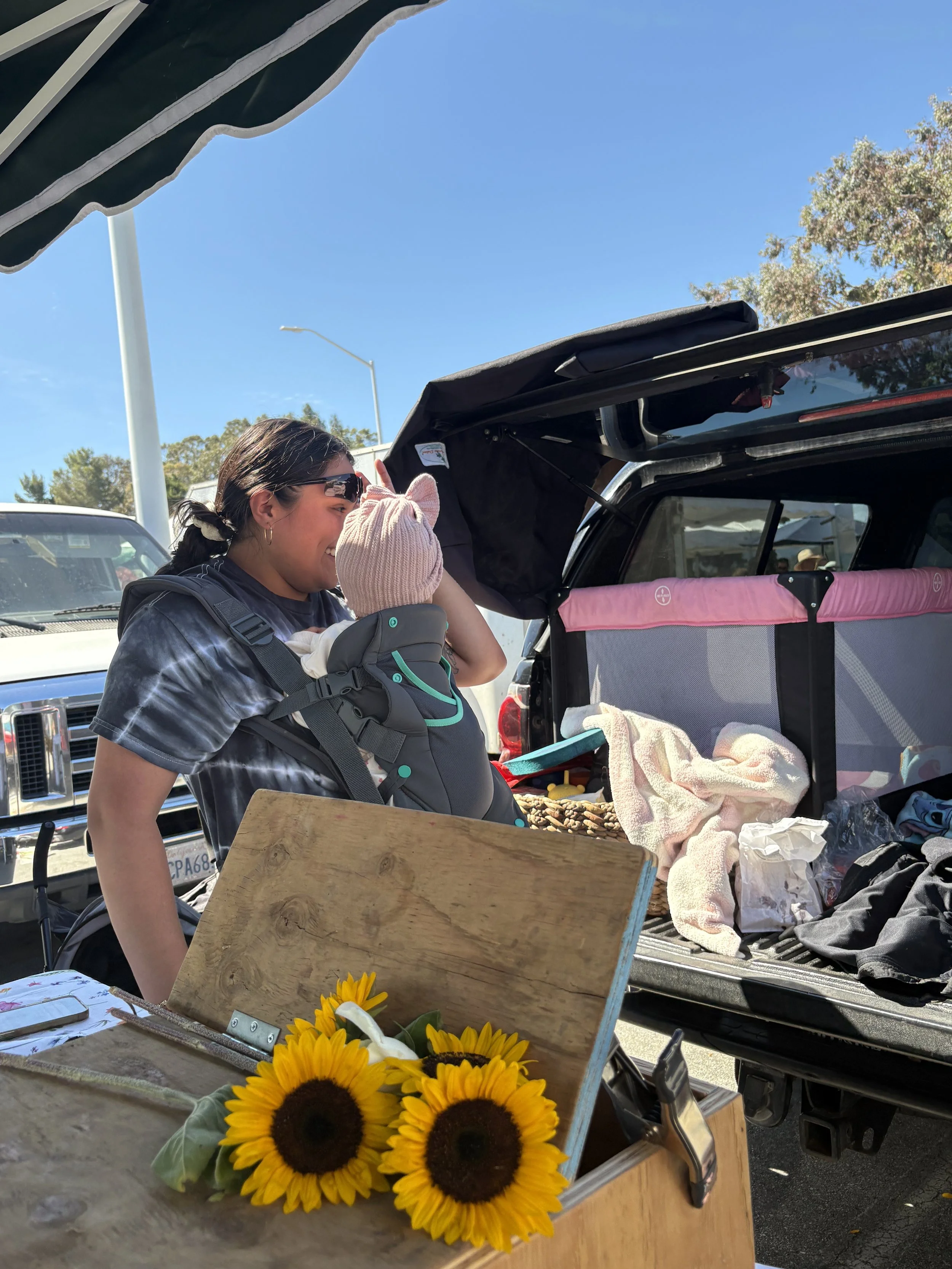 A woman wearing sunglasses and a backpack is smiling and talking to a small child in a carrier. They are at an outdoor market or event with parked cars and tents in the background. The open trunk of a vehicle contains a pink storage compartment, towe