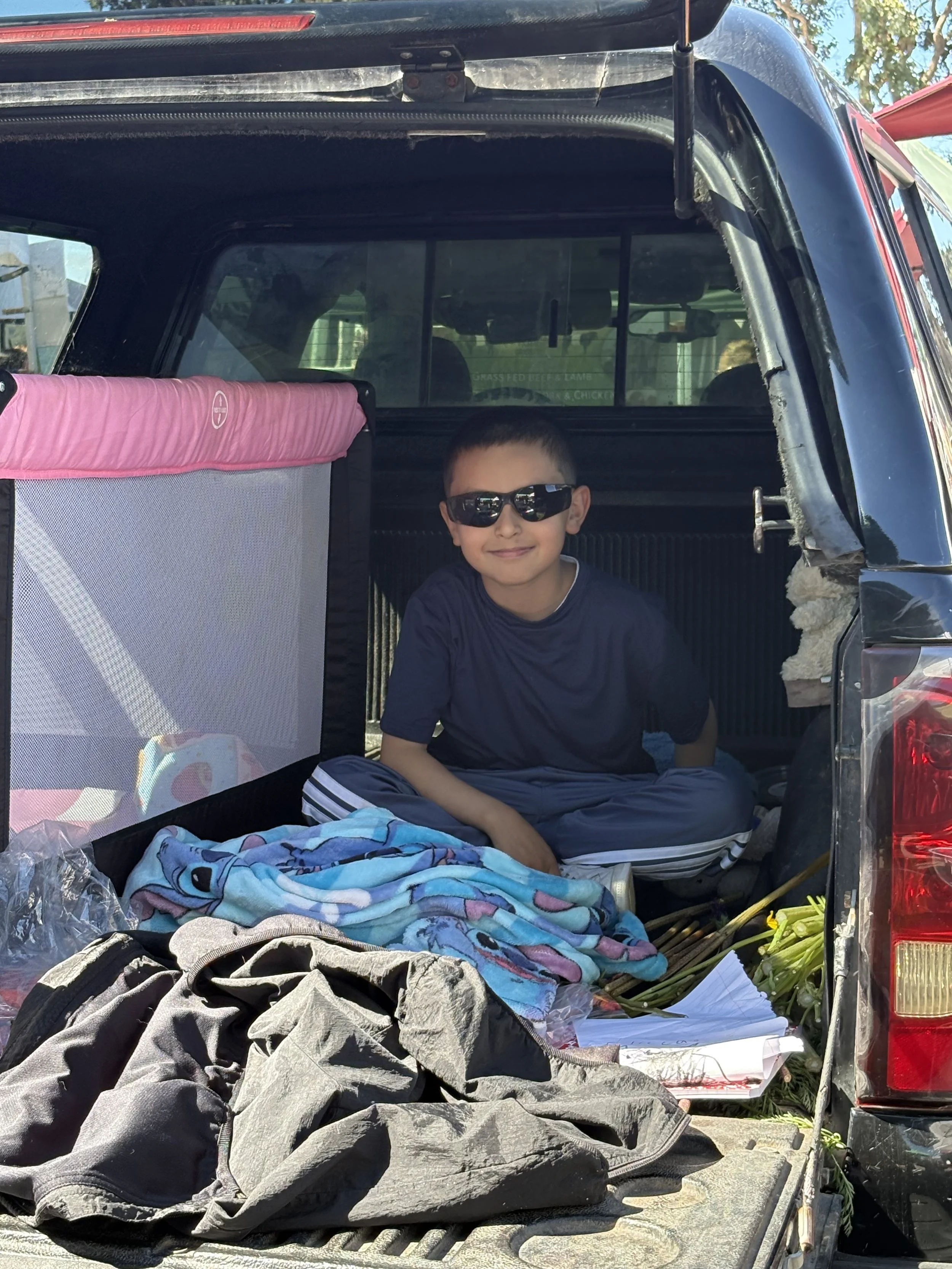 A young boy wearing sunglasses, sitting in the back of a pickup truck with various items including blankets and clothes, outdoors during daytime.