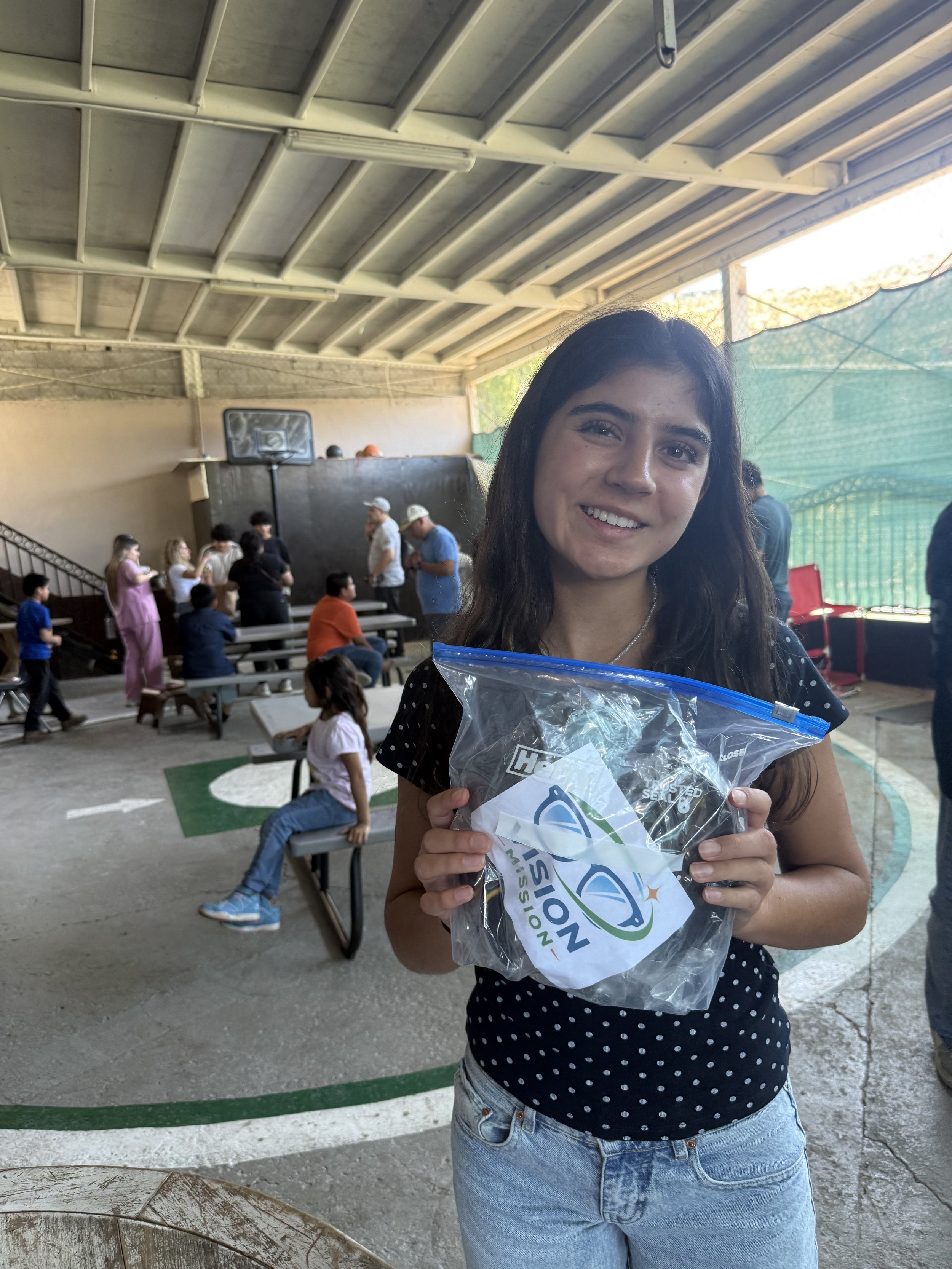 A young woman with dark hair smiling, holding a clear plastic bag with a logo, at an outdoor covered space with people in the background.