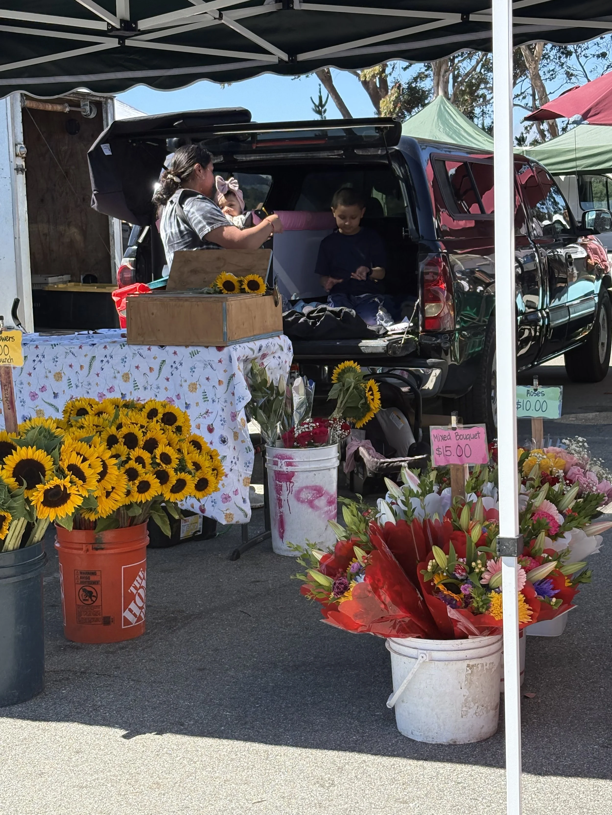 A flower stand at an outdoor market with various flowers such as sunflowers and mixed bouquets. The stand is shaded by tents, and a table with a floral cloth displays flowers. Buckets and signs showing prices are visible, with a woman and children in