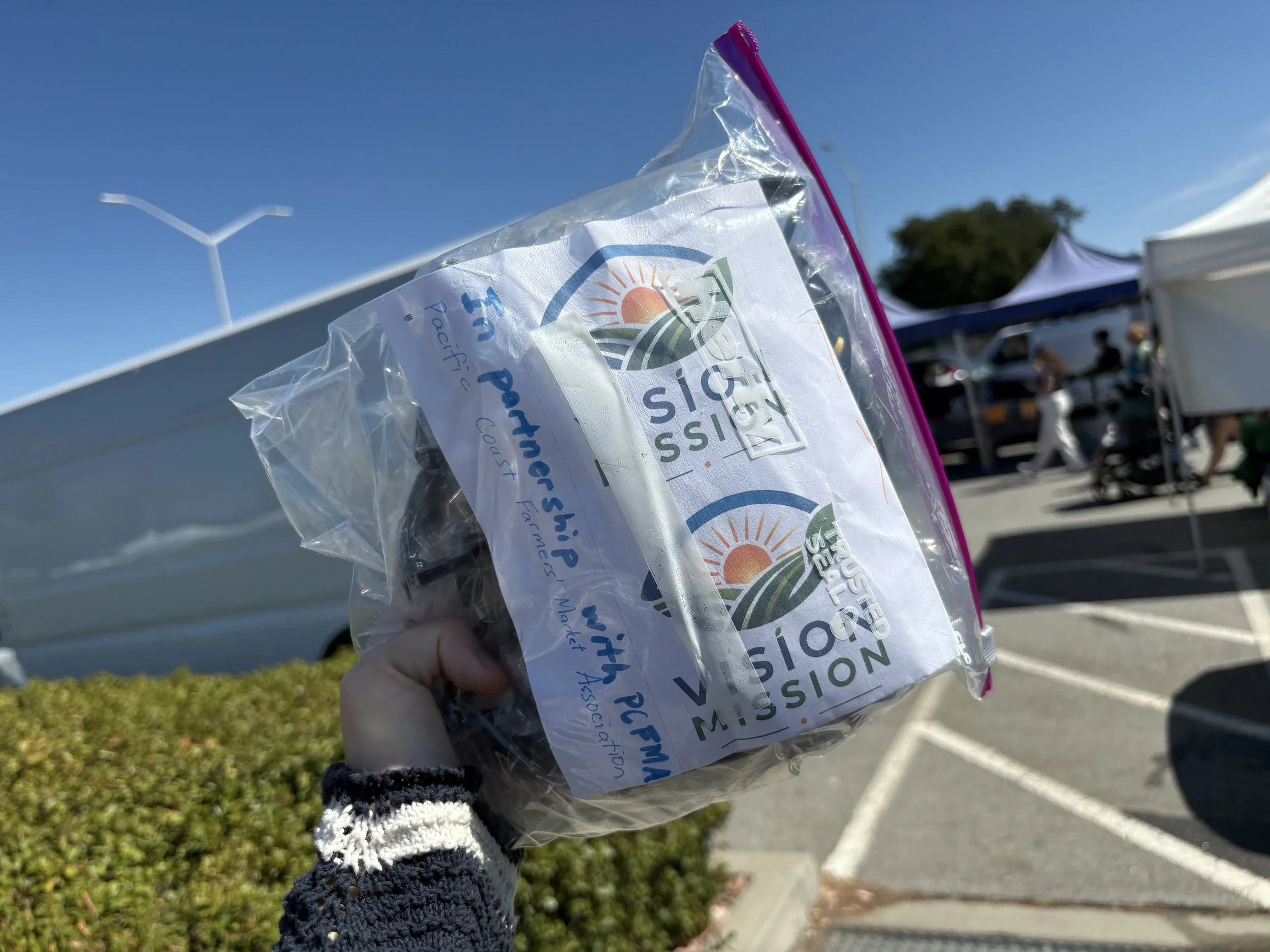 A person holding a plastic bag with a logo and text that reads "Mission
Fusion" and "Project" at an outdoor event. There are tents, people, and a clear blue sky in the background.