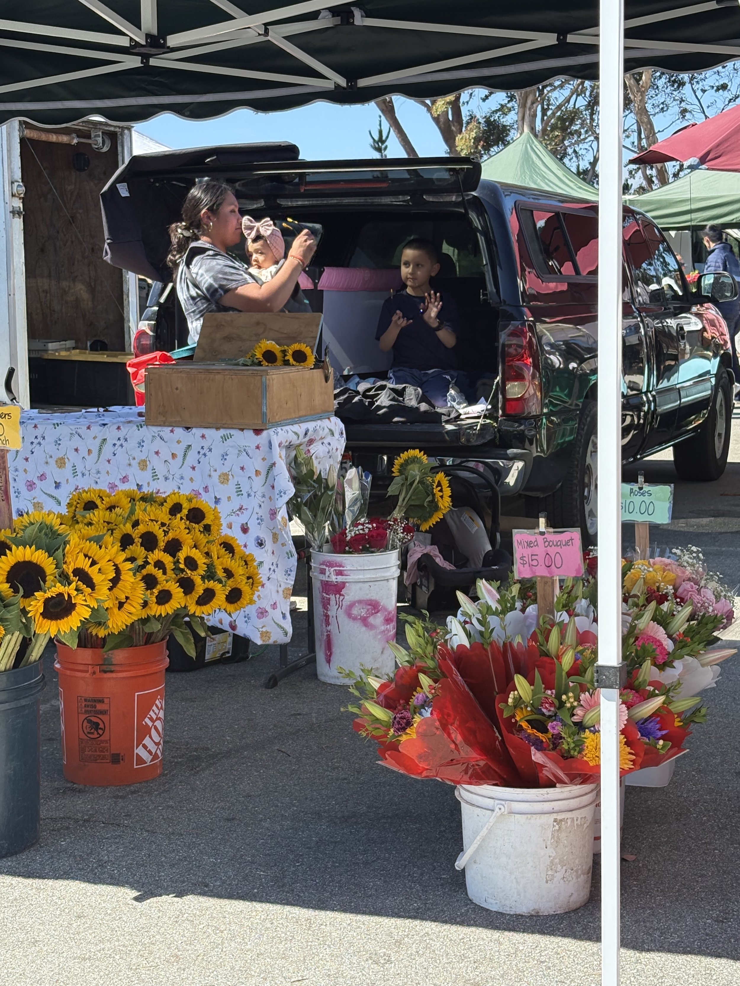 A flower vendor's booth at an outdoor market, displaying various colorful bouquets and sunflowers, with two children and a woman underneath a canopy.