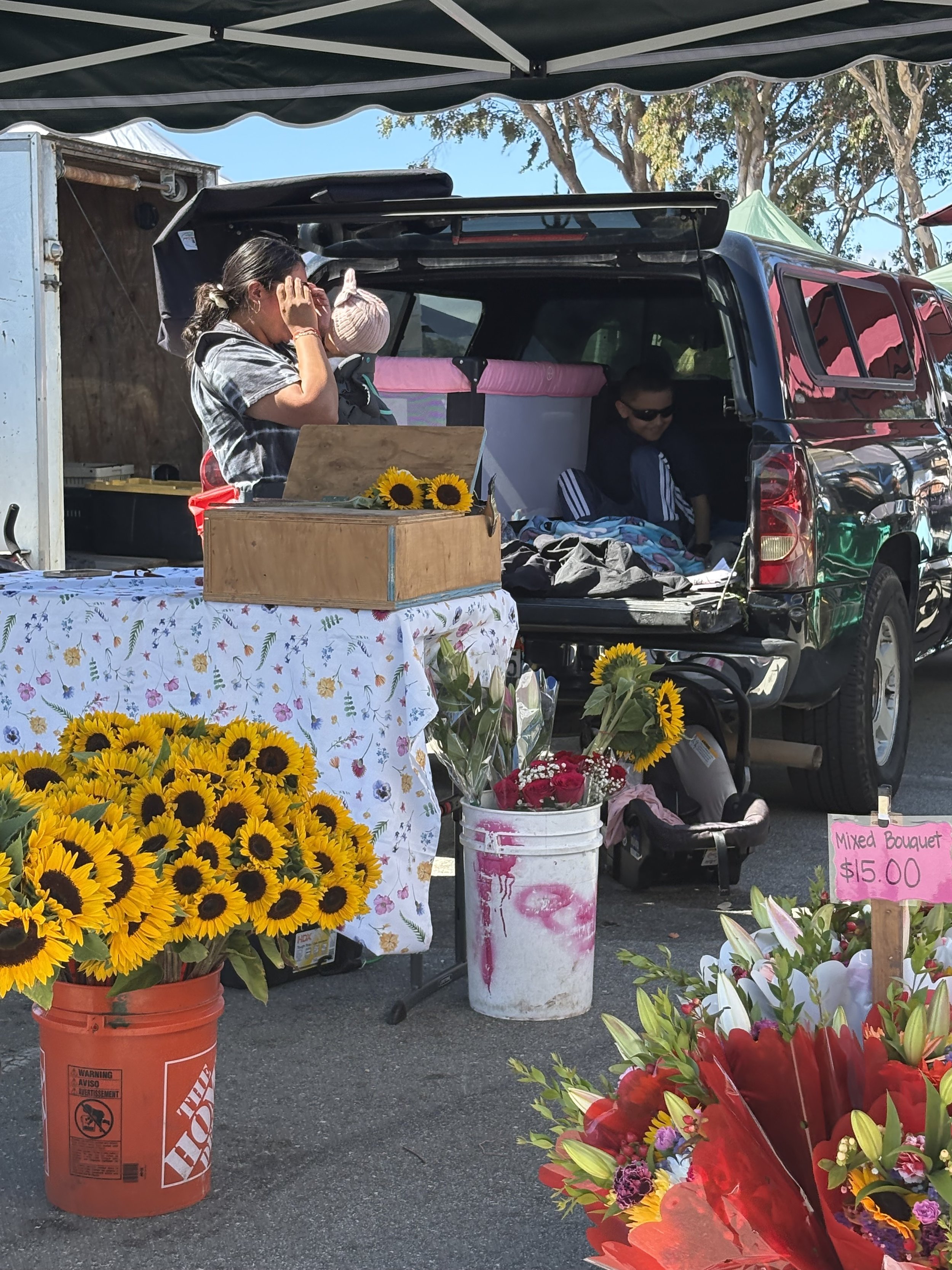 A flower stand at an outdoor market with sunflowers, mixed bouquets, and other flowers, a table with a floral tablecloth, and a sign indicating mixed bouquets for $15.00. There are people browsing, and a black vehicle is parked behind the stand.