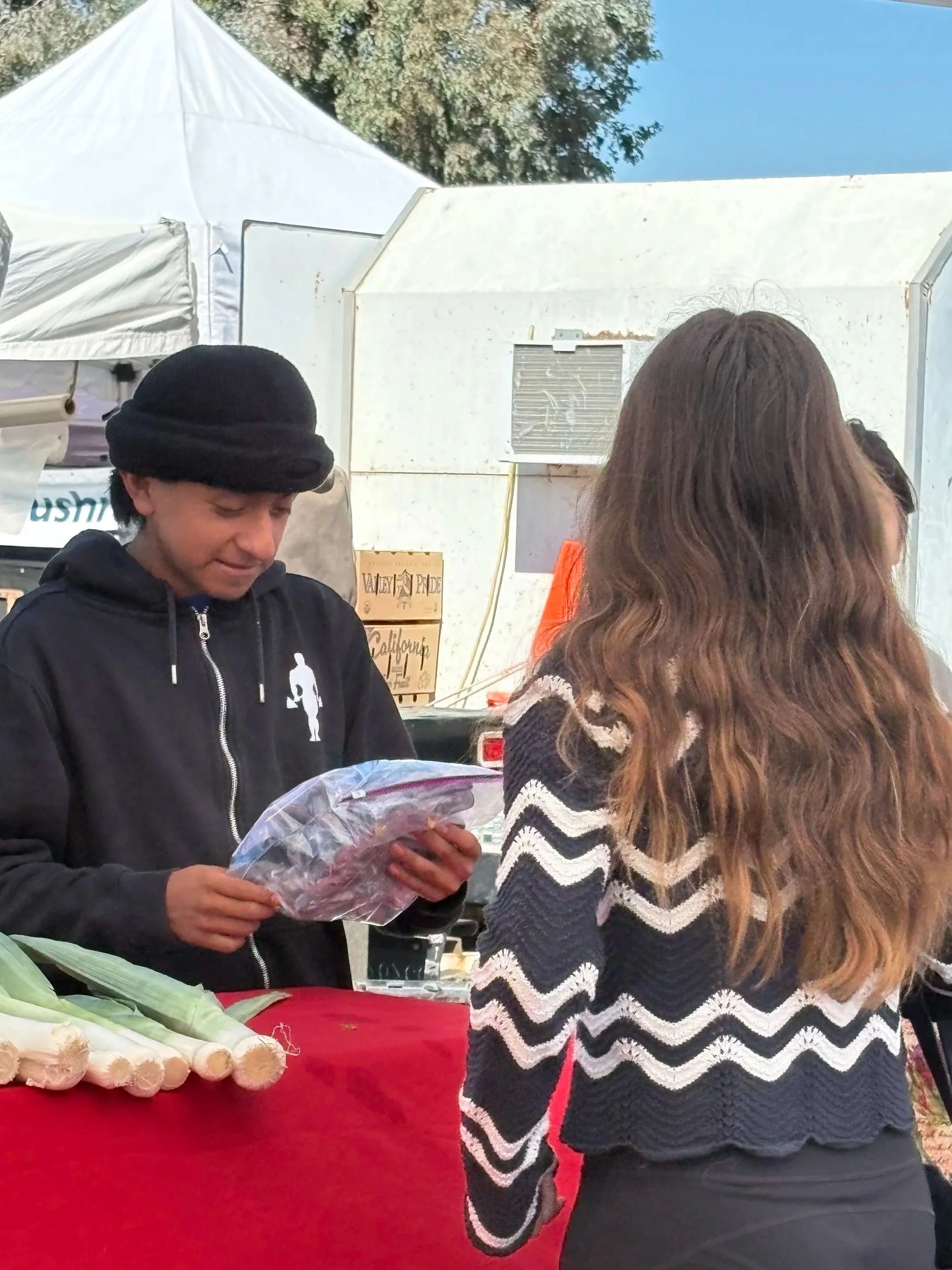 A man wearing a black hoodie and black hat is holding a plastic bag of meat at an outdoor market. The back of a woman with long, wavy brown hair and a black and white zigzag sweater is visible in front of him. There is a red tablecloth with green vegetables and garlic, and white tents and trees in the background.