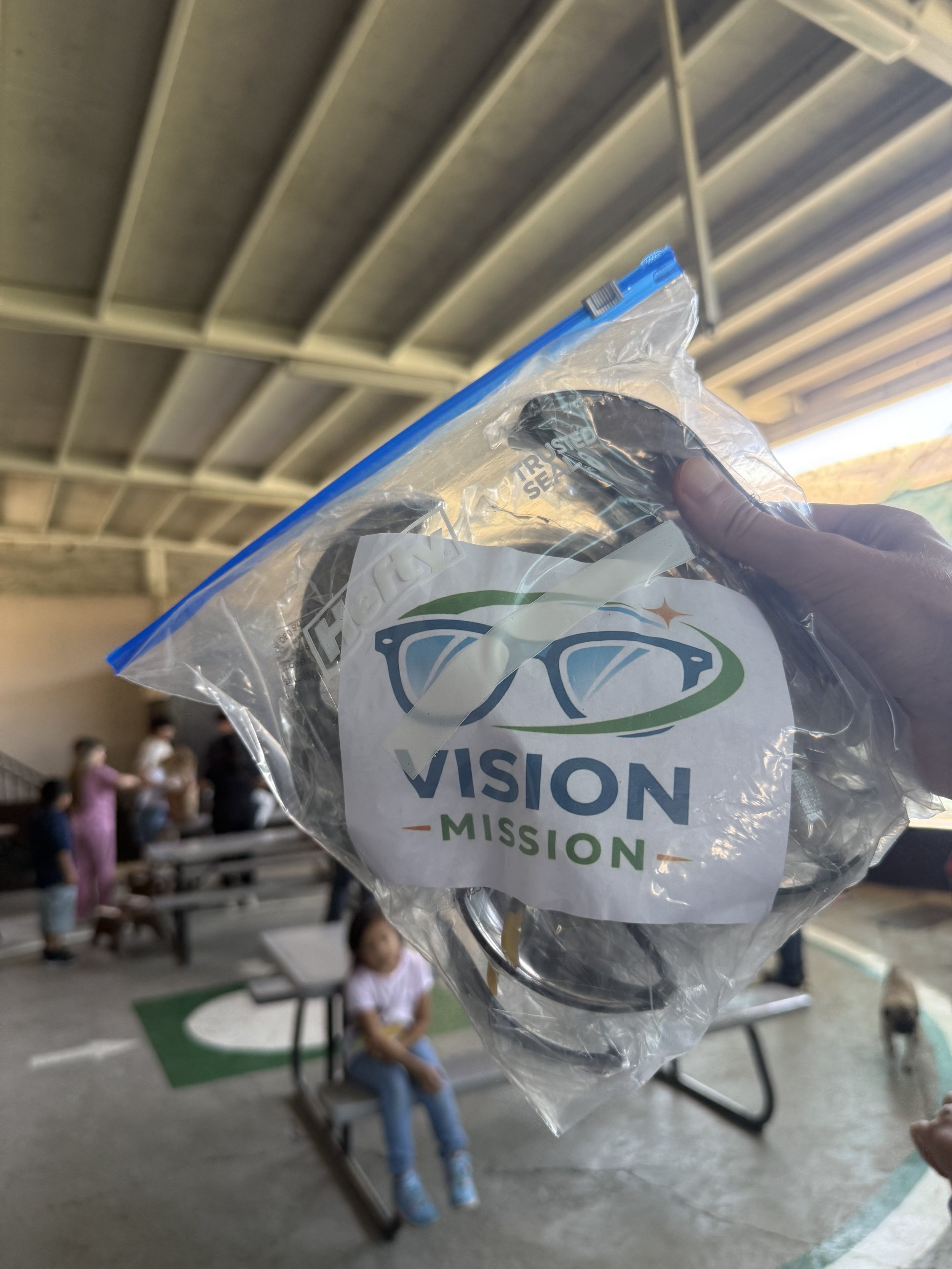 A hand holding a clear plastic bag with a black circular object and a label that reads 'VISION MISSION' at an indoor gathering, with children sitting on benches in the background.